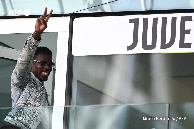 A man with dark skin and short, curly hair is seen waving from behind a glass panel. He wears stylish sunglasses and a patterned denim jacket. He is giving a peace sign with his right hand while having a broad smile on his face. The background features the word "JUVE" prominently displayed in bold black letters against a white area, indicating the Juventus Football Club. The setting suggests a sports environment, likely within a stadium, where fans and players might gather. The atmosphere appears joyful and optimistic, reflecting the man's positive demeanor amidst recent challenges.