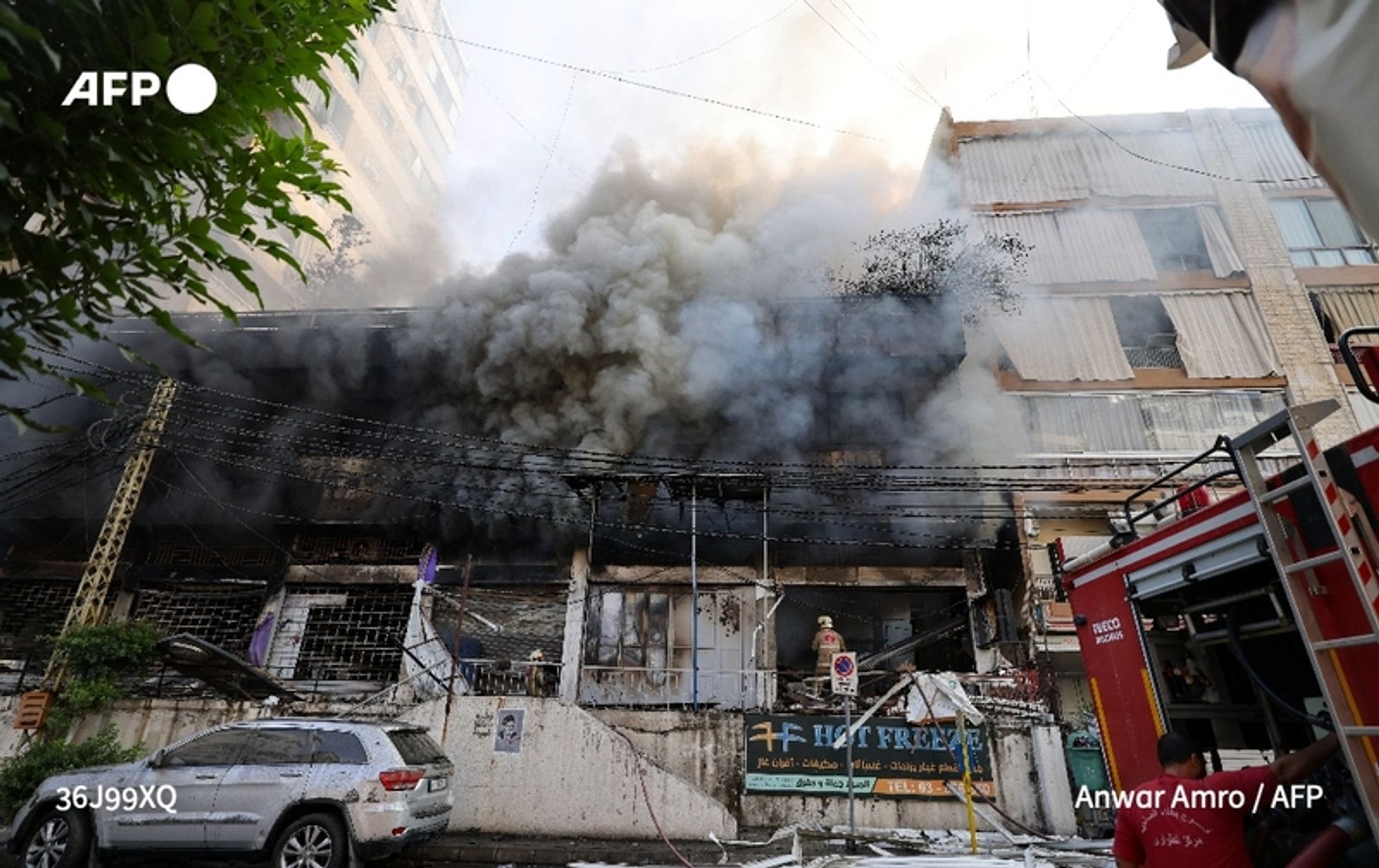 A scene depicting the aftermath of a violent event in Beirut, with a large building engulfed in thick, dark smoke billowing into the sky from the roof. The building appears damaged, with sections of its exterior showing signs of destruction. To the left, a white vehicle is parked on the street, partially visible beneath nearby foliage. In the foreground, a firefighter in a yellow uniform can be seen assessing the situation, positioned near the entrance of the building. Firefighting equipment is visible, indicating ongoing efforts to control the blaze. The overall atmosphere conveys a sense of urgency and chaos in the wake of the strikes targeting Hezbollah, with smoke rising ominously against the backdrop of a clear sky.