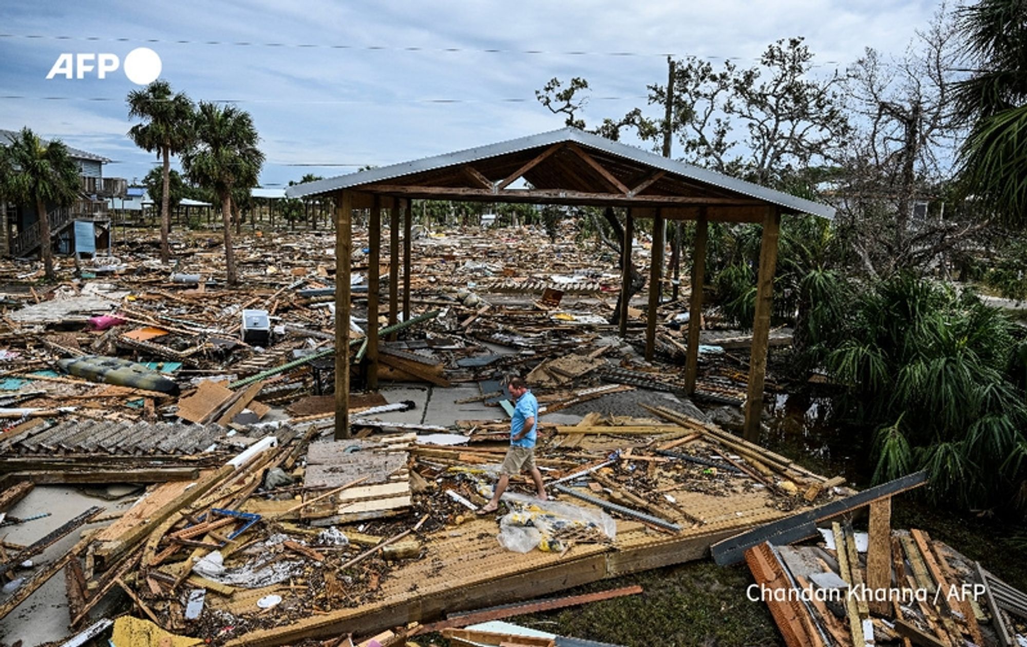 A wide-angle view of a devastated landscape following a powerful storm, likely caused by climate change. In the foreground, a person wearing a light blue shirt is walking through a chaotic scene of destruction, surrounded by piles of debris, broken wooden structures, and scattered roofing materials. The ground is littered with planks, fragments of buildings, and vegetation remnants. In the background, remnants of palm trees can be seen against a cloudy sky, indicating a tropical setting. An open-air structure with a roof and wooden beams stands prominently, partially intact amidst the destruction. The overall atmosphere conveys a sense of loss and the aftermath of a cyclone, emphasizing the amplified impact of climate change on weather events.