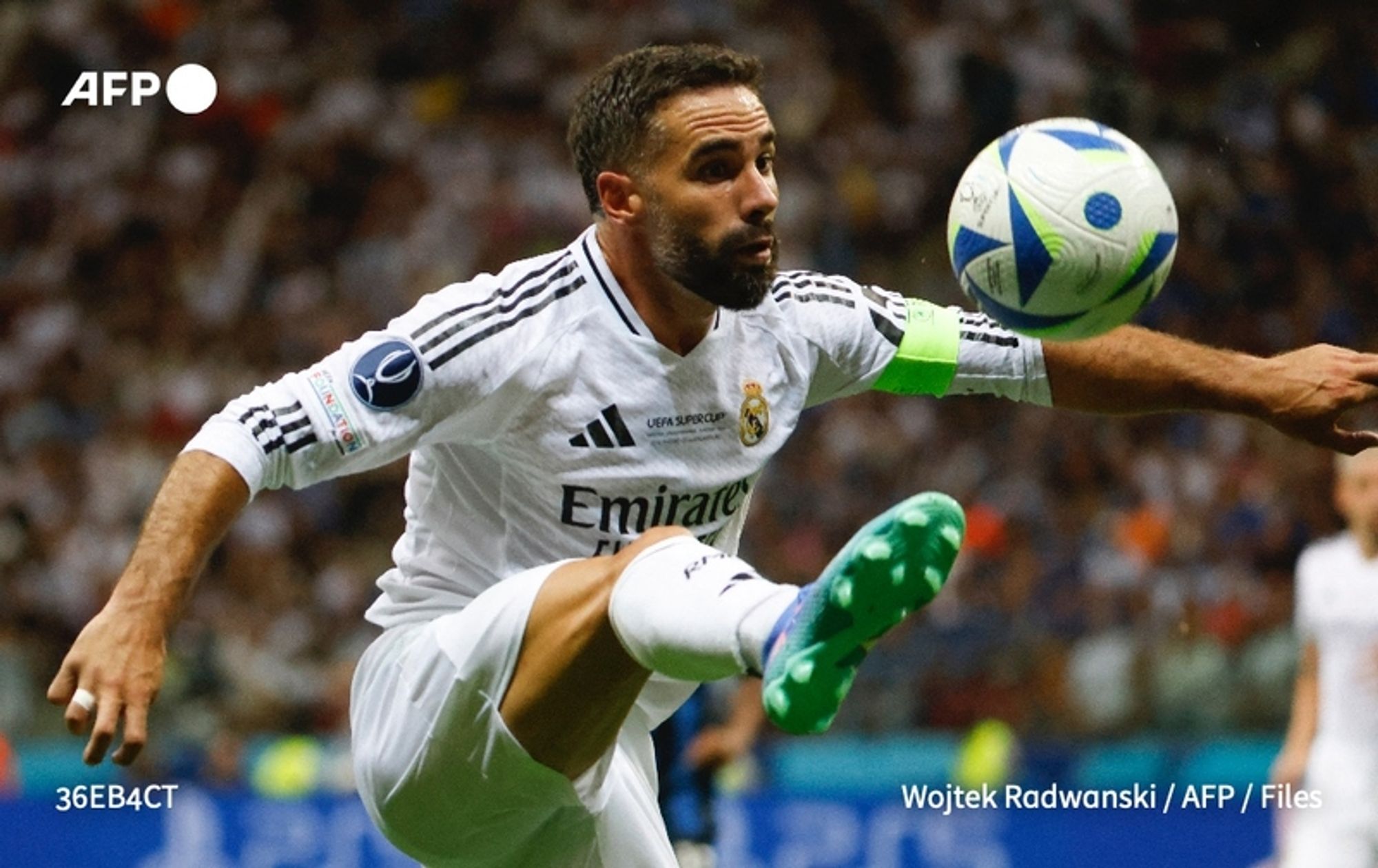 A male soccer player in action during a match, wearing a white uniform with black and gold accents, representing Real Madrid. The player, who has short dark hair and a trimmed beard, is captured mid-stride as he kicks a soccer ball with his right foot. He is wearing a captain's armband on his left arm and has an intense focus on the ball. The background is a blurred stadium filled with spectators, suggesting a vibrant game atmosphere. The player's body is slightly turned, and his expression reflects concentration and determination. The soccer ball is patterned with blue and green, prominently displayed in the foreground.