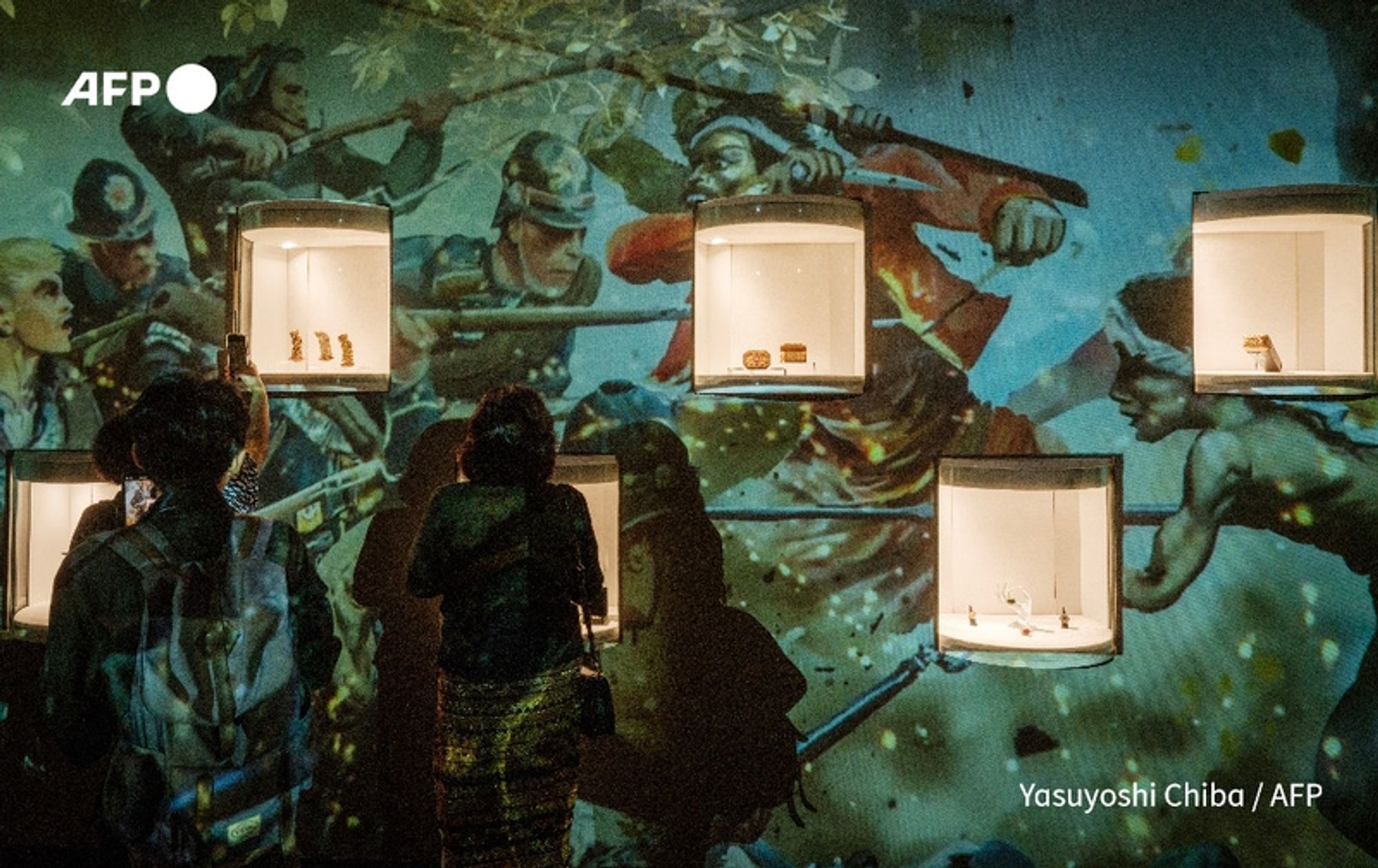 A group of visitors is seen in a dimly lit exhibition space at the National Museum of Indonesia in Jakarta. They are facing multiple display cases illuminated against a vibrant backdrop. The background features a large mural depicting historical scenes with figures engaged in conflict, suggesting a narrative of struggle or battle. The display cases are mounted on the wall and showcase intricate jewellery artifacts from the repatriated Lombok collection, alongside a recently returned statue of Brahma. The visitors, dressed in dark attire, are observing the artifacts intently, highlighting their cultural significance. The atmosphere conveys a sense of reverence and curiosity about the repatriated items and their historical context.