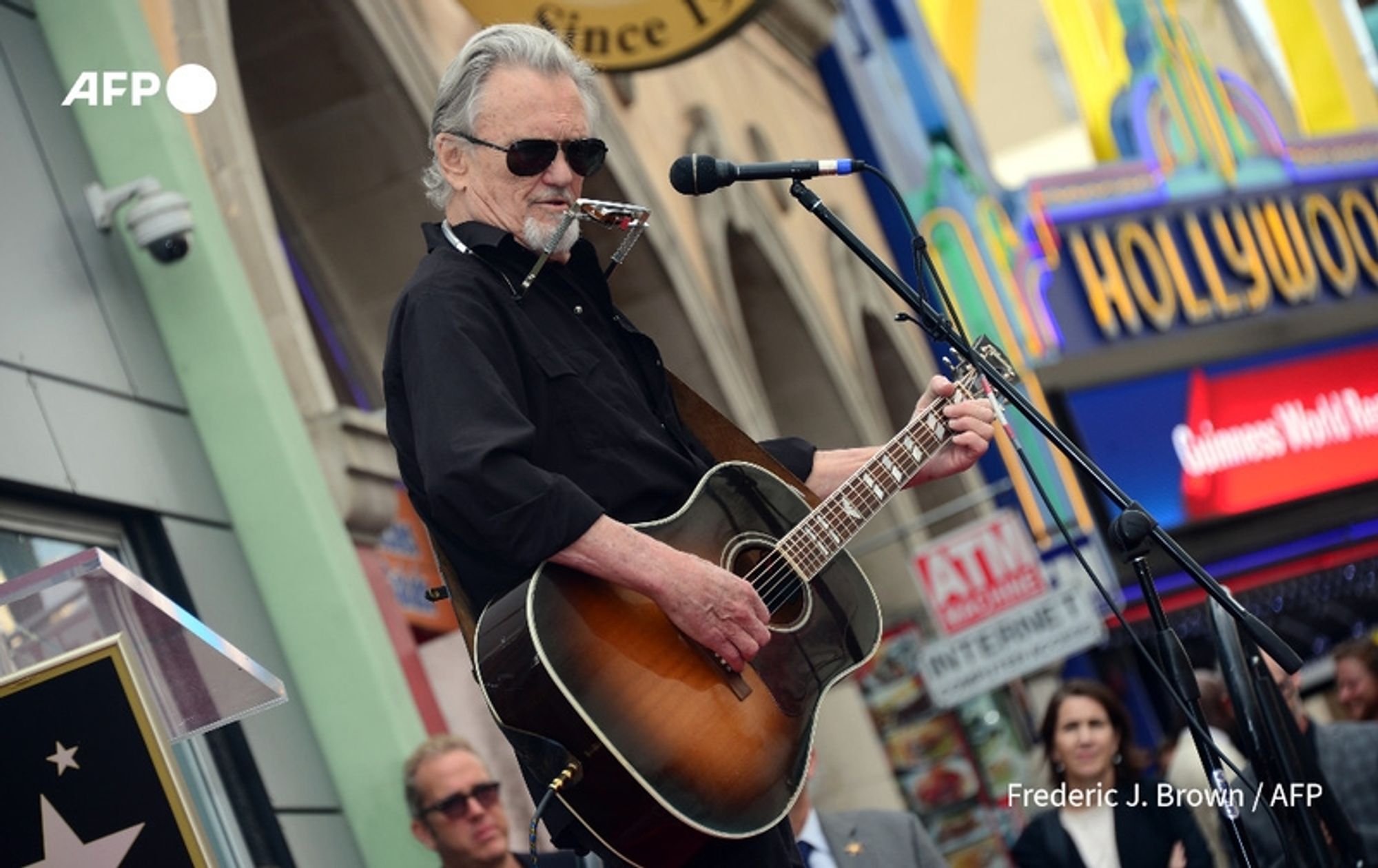 A 88-year-old man stands on a stage, holding an acoustic guitar, wearing sunglasses and a black button-up shirt with the sleeves rolled up. He has gray hair, slicked back, and is partially turned towards a microphone on a stand. A harmonica is clipped to his shirt, adding to his musician's appearance. In the background, colorful signage displays the word "HOLLYWOOD," surrounded by various shop fronts. Some spectators are visible, listening intently, with one person, a woman in a cream-colored jacket, standing prominently in the foreground, while others appear in soft focus. The setting suggests an outdoor event, likely a tribute or celebration, reflecting the individual’s contributions to music and film.