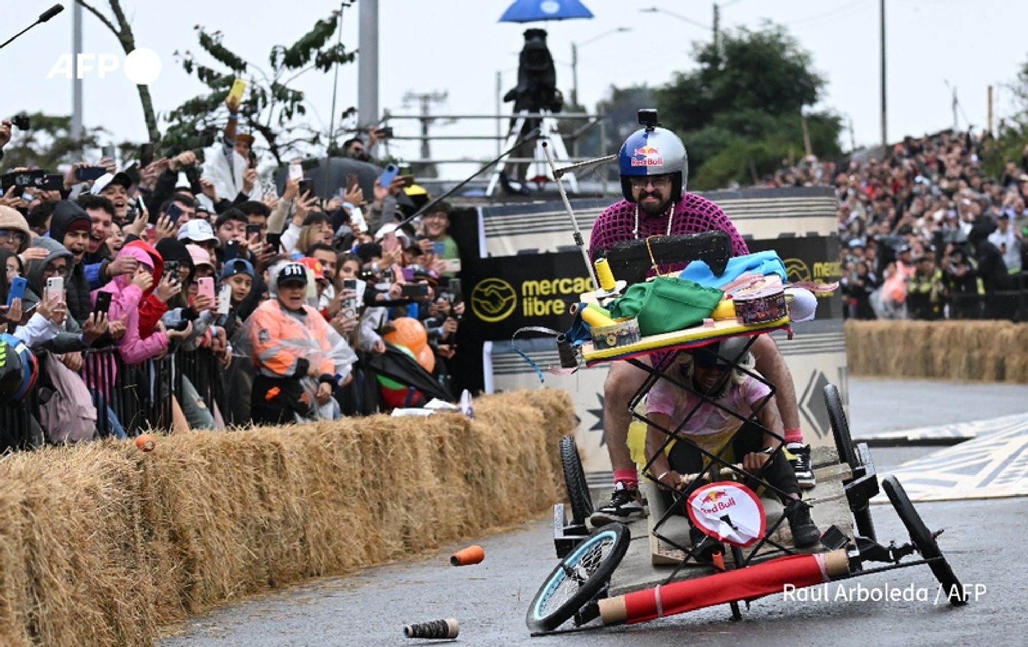 A lively scene from the annual "Balineras" race in Bogotá, Colombia, captures participants competing on homemade carts. In the foreground, a man rides a colorful, improvised cart that is loaded with various items, including cushions and a container, suggesting a whimsical design. He wears a helmet adorned with a camera, and his expression conveys excitement. Directly beneath him, another participant is seen lying flat on the cart, contributing to the dynamic action. Spectators line the side, many raising their phones to capture the moment, their expressions a mix of thrill and amusement. The atmosphere is vibrant, with greenery, urban infrastructure, and eager crowds in the background, emphasizing the festive spirit of the event. Hay bales are positioned along the track, and a large banner is visible in the back, adding to the colorful and energetic ambiance of this unique obstacle course race.