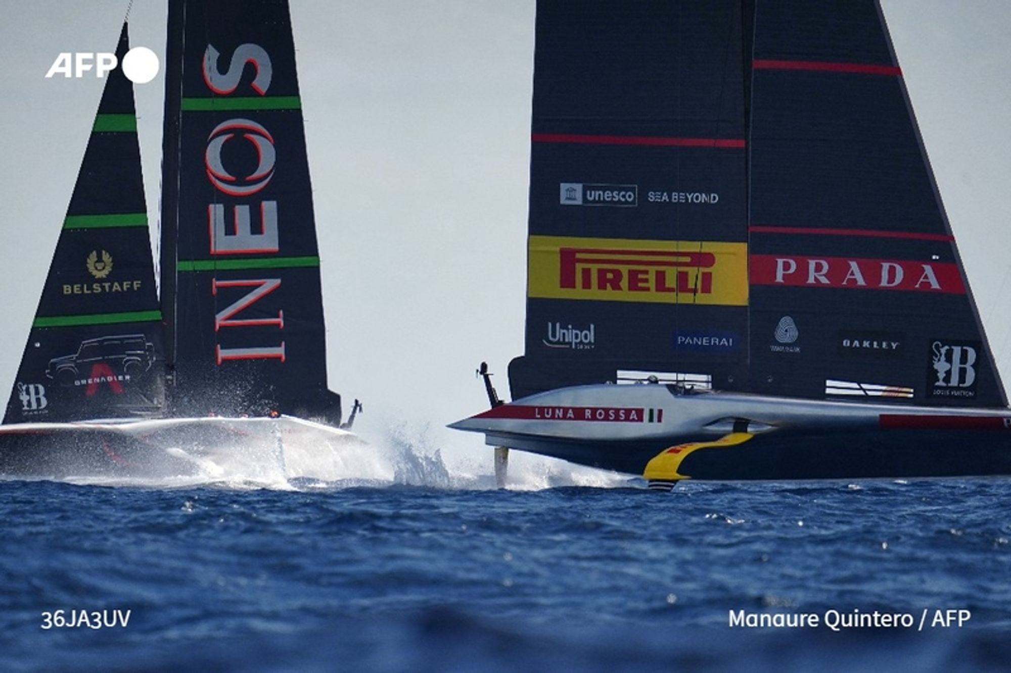 Two high-tech racing yachts are depicted competing in a thrilling match on the shimmering blue waters off the coast of Barcelona. The yacht on the left, sporting a dark hull, is emblazoned with the name "INEOS" in large white letters along its sail, which also features green and red stripes, and brand logos, including Belstaff. The yacht on the right, identified as "LUNA ROSSA," displays a sleek white hull with a colorful design and several sponsor logos on its black sails, including Pirelli and Unipol. The scene captures the dynamic moment of the race, with both boats cutting through the water, creating splashes as they sail close to one another, showcasing the excitement of the Louis Vuitton Cup qualifying event. The background reveals a bright sky, enhancing the visual impact of this competitive racing moment.