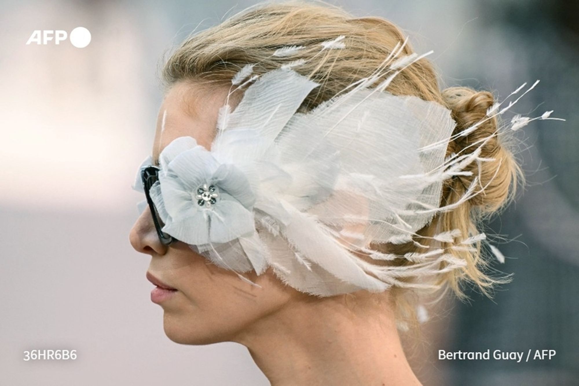 A woman is depicted in profile, showcasing an elaborate headpiece that resembles a birdcage theme, reflective of the Chanel fashion show. Her blonde hair is elegantly styled in a low bun, with strands gently falling around her face. The headpiece features delicate white feathers and tulle, adorned with a large white flower at the front, adding a whimsical touch. She wears dark rectangular sunglasses that contrast with her light features, and her expression is serene. The background is softly blurred, emphasizing her and the ornate details of her headwear, aligning with the luxurious aesthetic of the Chanel show held at the Grand Palais.