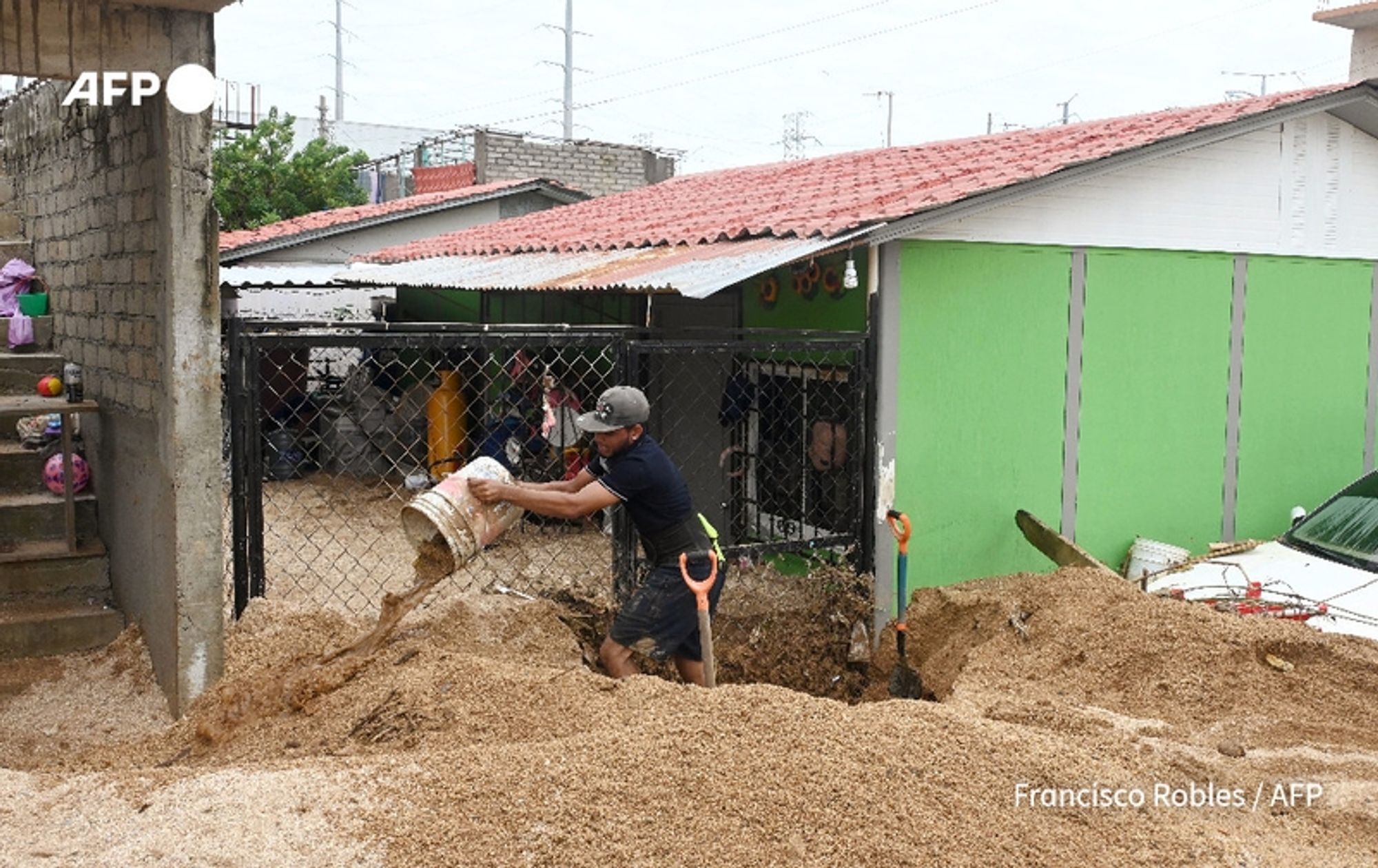 A man wearing a black cap and a dark shirt is shoveling sand or debris from a large pile into a bucket. He is positioned outside a fenced area in a residential neighborhood that shows signs of recent flooding, likely due to Hurricane John. A partially visible house with a red tiled roof and brightly painted green walls can be seen behind him. The environment appears affected by the storm, with muddy ground and visible water damage. In the background, several other houses are visible, with various colors and structures hinting at a close-knit community. A figure can be seen inside the fenced area, watching the man work. The overall scene conveys the aftermath of a natural disaster, highlighting efforts to clean up and recover from the flooding.