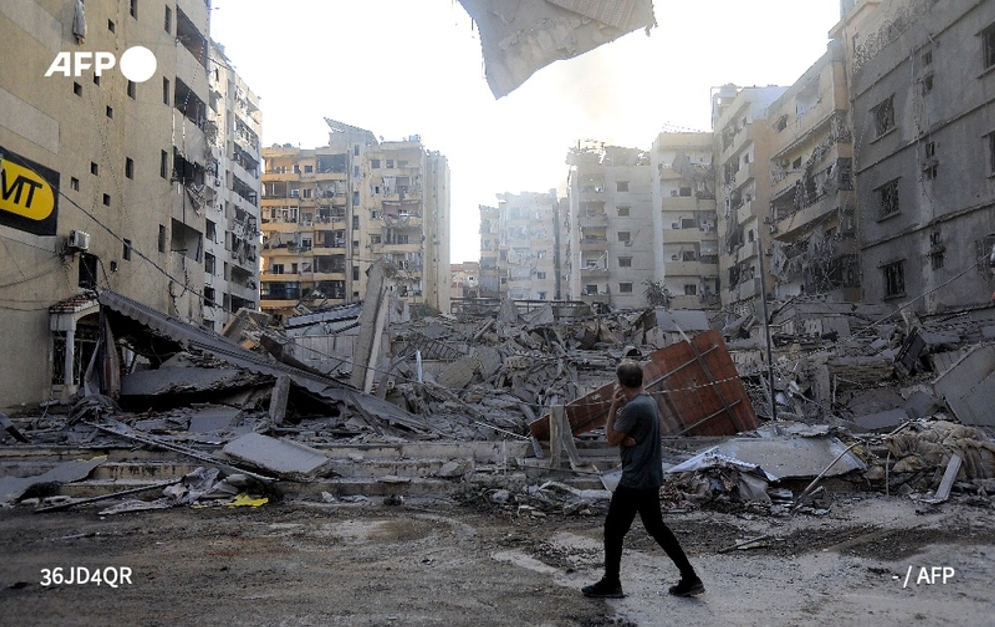 A person is walking through a devastated urban landscape in Beirut. The scene shows collapsed buildings and extensive rubble scattered across the ground, highlighting the destruction in the area. Dust and smoke linger in the air, suggesting recent violence. The individual, seen from behind, wears a casual grey shirt and appears contemplative as they navigate through the debris. Surrounding buildings are partially standing, some with broken windows and damaged facades, indicative of recent intense strikes. The atmosphere conveys a sense of devastation and loss, underscoring the impact of ongoing conflict.