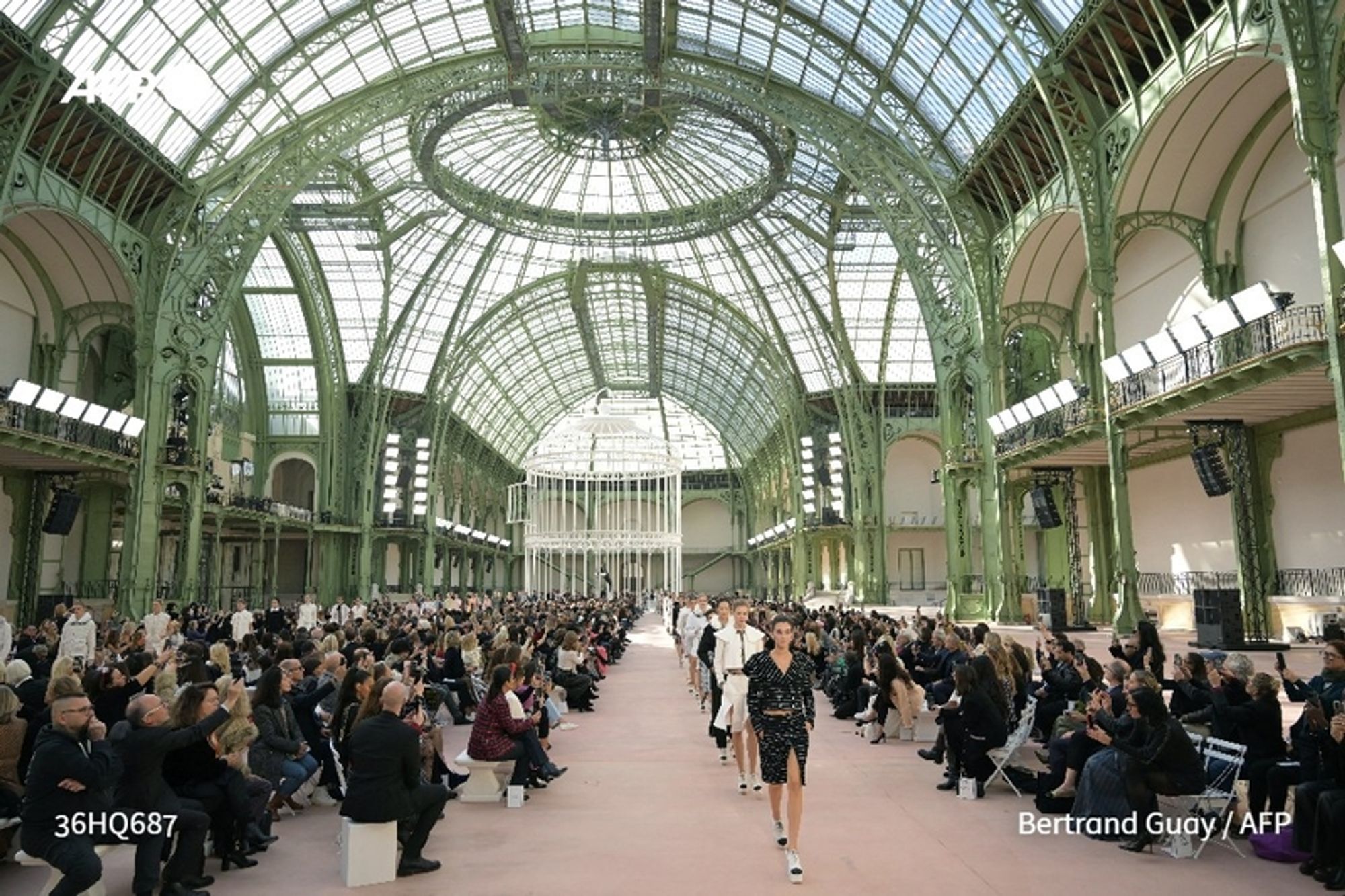 A wide-angle view of the Grand Palais in Paris during a Chanel fashion show. The venue features a stunning glass and metal structure with a high arched roof adorned with intricate green detailing, providing sunlight to the interior. The runway is centered, leading towards a minimalist white installation, enhancing the birdcage theme of the event. A diverse audience is seated on either side of the runway, attentively watching the models who are walking the runway in stylish outfits, reminiscent of Chanel's past glories. The ambiance combines elegance and excitement, capturing the haute couture essence of the show. The floor is covered in pale pink material, contrasting with the greenery of the architecture and the black, white, and patterned ensembles of the models.