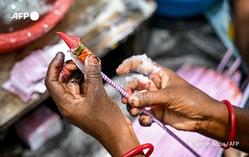 A close-up view of two hands working in a fireworks factory in India, preparing items for the Diwali festival. One hand holds a cone-shaped firecracker wrapped in red paper, displaying a colorful label. The fingers are stained, indicating hands-on work with fireworks. In the background, multiple thin, pink paper tubes are visible, suggestive of assembly line production. The workspace is cluttered, with out-of-focus items hinting at an industrious environment. The overall scene conveys a sense of urgency and craftsmanship as workers hasten to meet high demands for the festival of lights.