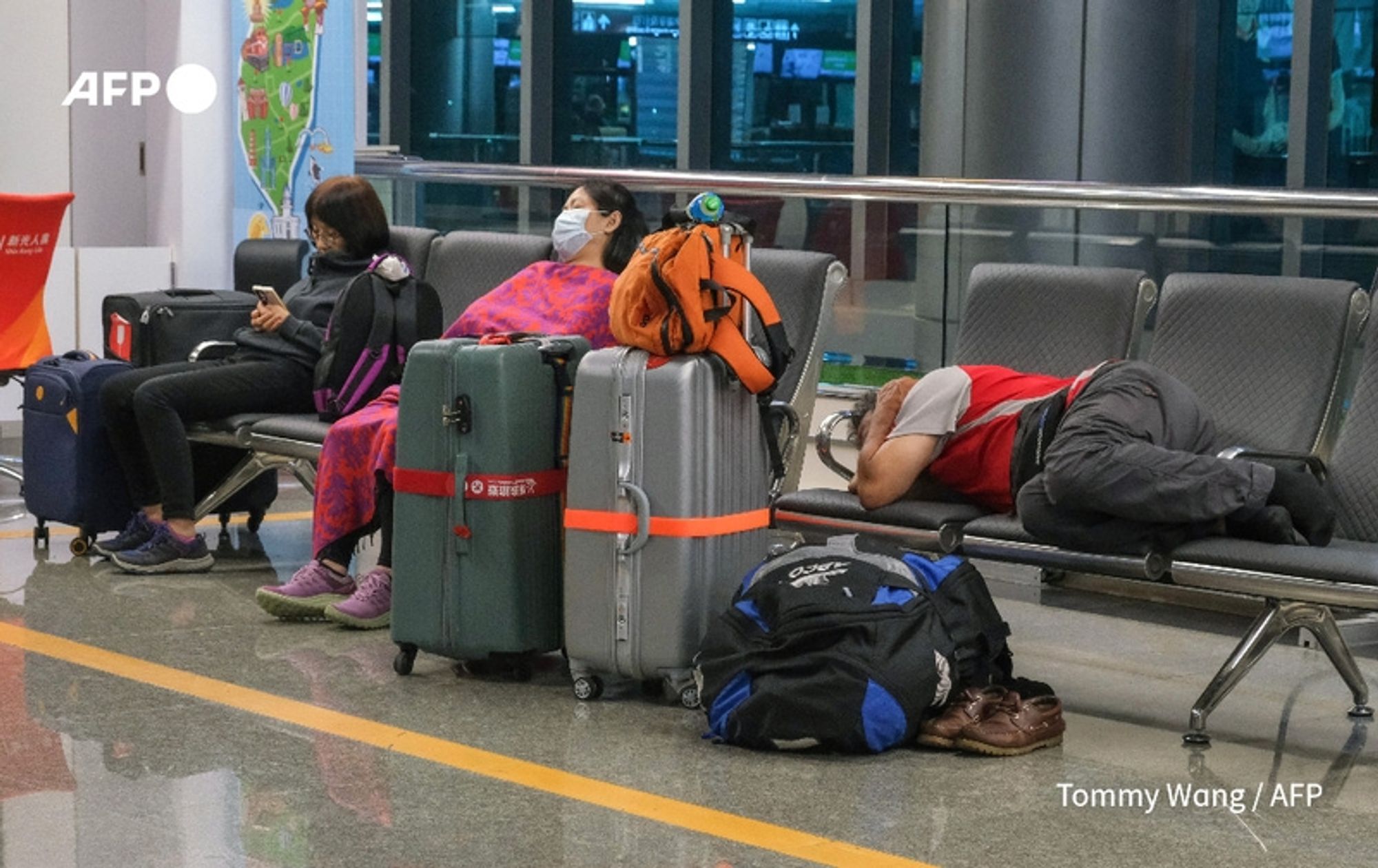 A scene inside an airport terminal during Typhoon Krathon's approach in Taiwan. Three travelers are seated on metal benches, each accompanied by various pieces of luggage. On the left, a woman in a black top and pink pants is sitting with her feet up, looking at her phone. She is wearing a mask. Next to her, another woman is reclining against her suitcase, dressed in a bright pink and orange patterned outfit, also wearing a mask, with a backpack resting beside her. On the right, a man, dressed in a red and gray shirt, is lying back on the bench with his head down, appearing to be asleep. Luggage scattered around includes three large suitcases in neutral colors, one with an orange stripe, and a black and blue backpack. The terminal features large windows, displaying a dimly lit atmosphere reflecting the urgency of the situation, while a red sign and airport information can be seen in the background.