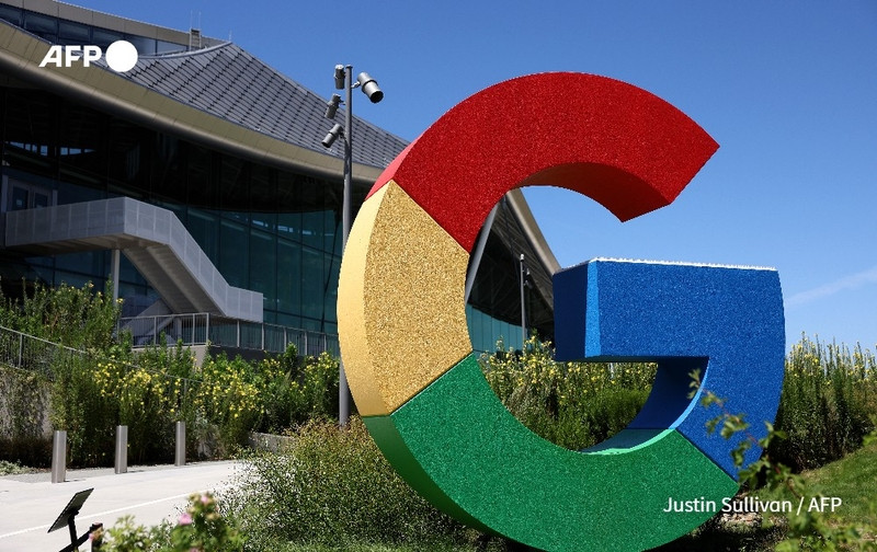 A vibrant three-dimensional sculpture of the Google logo is prominently displayed in the foreground. The logo consists of a large, rounded letter "G" made up of four colorful segments: red at the top, yellow on the right, green at the bottom, and blue on the left. Behind the sculpture, there is a glass-fronted modern building with a sloped roof, suggesting a contemporary architectural style. Lush greenery with flowering plants fills the area around the logo, contributing to a bright and inviting atmosphere. The sky is clear and sunny, enhancing the overall vibrant colors of the scene. The image captures a moment in time at Google's campus, symbolizing innovation and technological advancement. In the corner, there is a small logo reading "AFP," along with the name "Justin Sullivan" indicating the photographer.