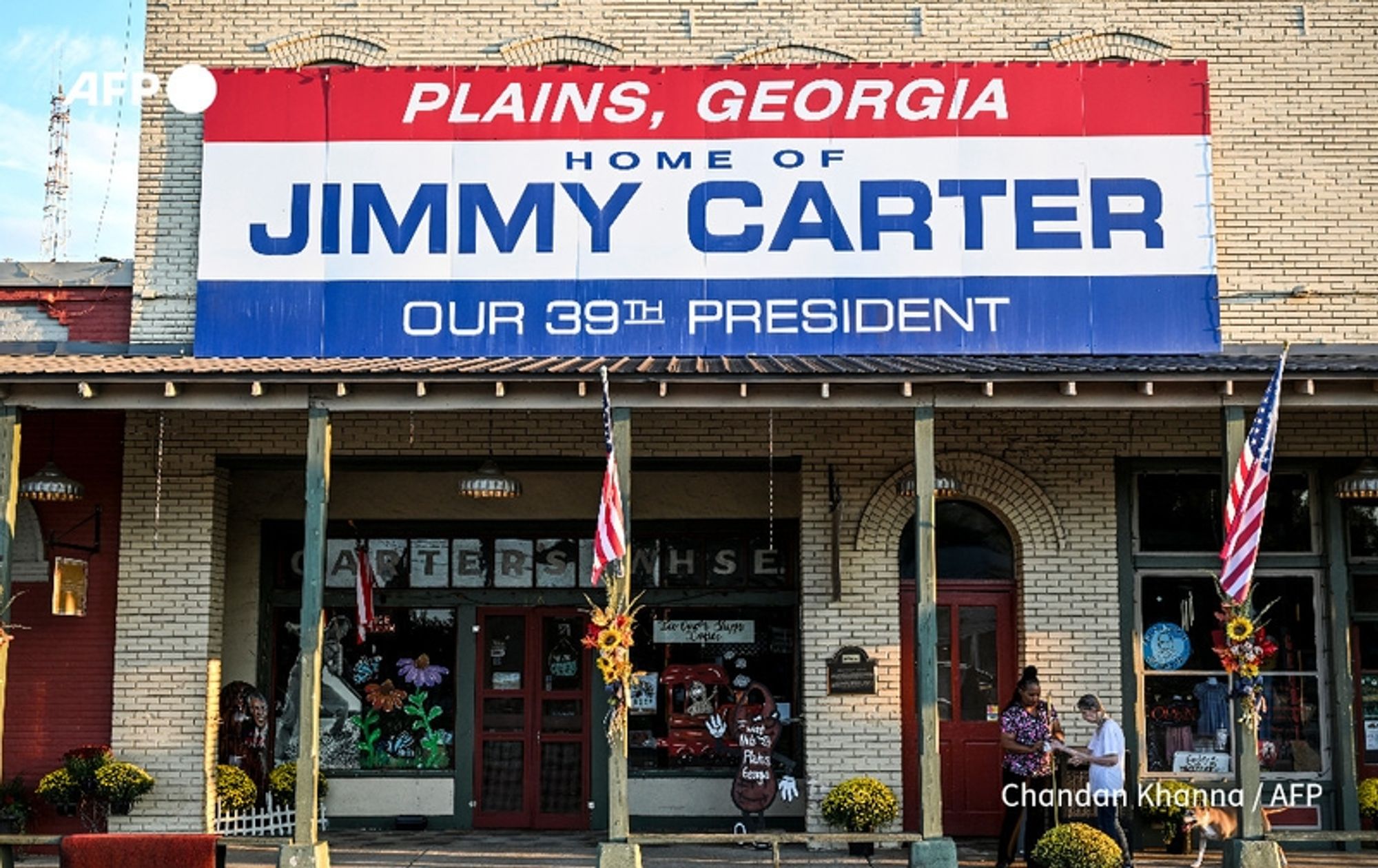 A colorful storefront sign prominently displays the words "PLAINS, GEORGIA" and "HOME OF JIMMY CARTER" in bold lettering. Below, it states "OUR 39th PRESIDENT." The sign is set against a light-colored brick building. In front of the entrance, two people are visible, engaging with the environment. Vibrant floral decorations and potted plants adorn the entryway, while American flags hang from the awning, adding to the patriotic ambiance. The scene captures the essence of Plains, Georgia, as it prepares to celebrate the centennial of Jimmy Carter, highlighting his significance as a beloved local figure.