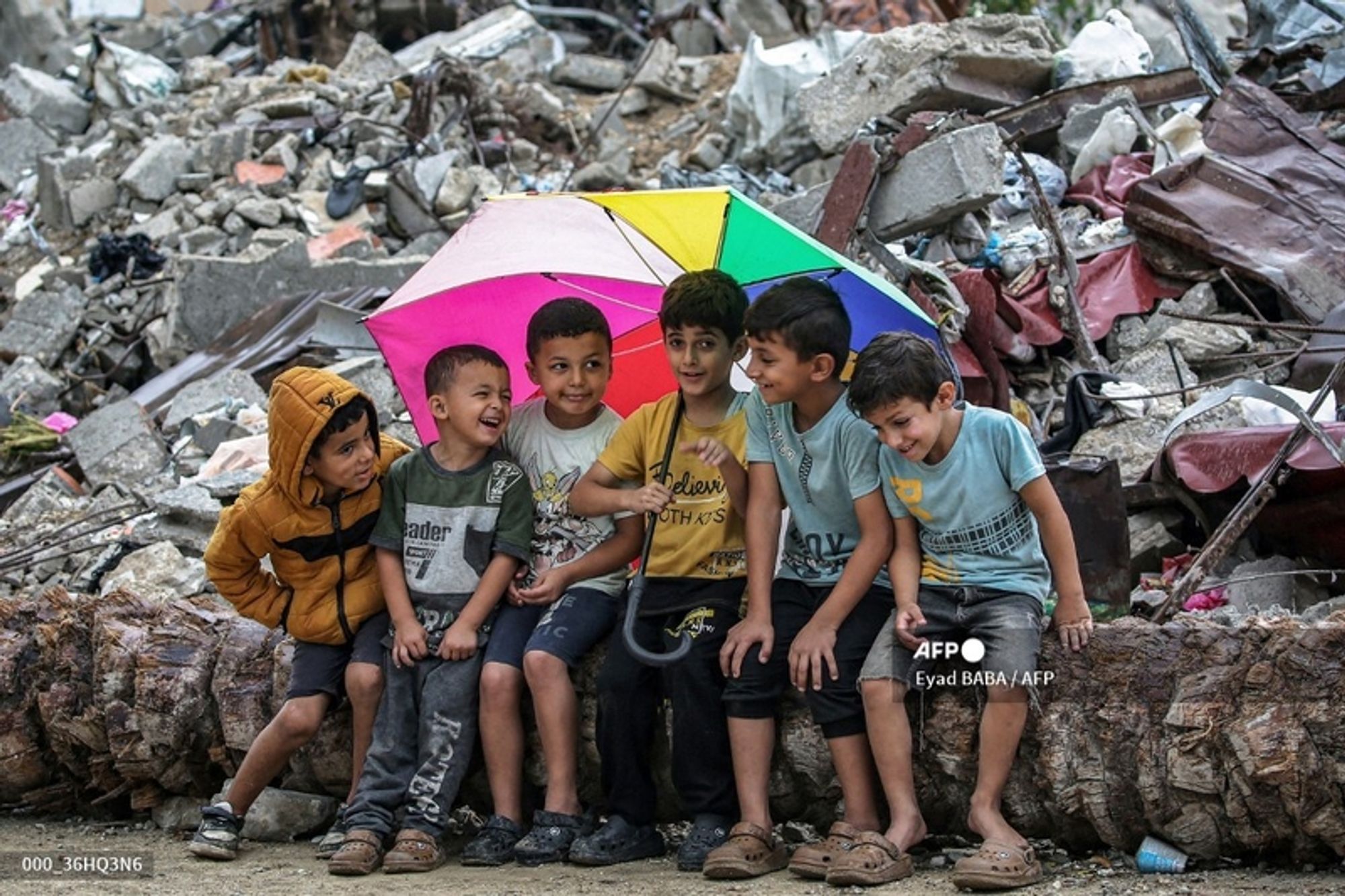 A group of six children sitting together in front of a pile of rubble and debris, likely from a damaged building or structure. The children are gathered under a large, colorful umbrella displaying a rainbow pattern, which provides a splash of color amid the gray surroundings. 

The child on the far left is wearing a brown hoodie with dark shorts and is looking at another child beside him, who is smiling broadly. The second child is wearing a T-shirt with a green design and is seated next to a boy in a yellow T-shirt. The latter is also smiling as he engages with the others. Next to him, another child in a light blue T-shirt appears to be sharing a joke or a moment of laughter with the group. To the far right, a child in light-colored shorts is looking down while smiling. 

The background features a chaotic scene of concrete and metal debris, highlighting the challenging environment in which the children are playing. The expressions on the children's faces convey joy and camaraderie despite their surroundings, suggesting a moment of happiness amidst adversity. The image is captured in natural light, enhancing the vivid colors of the umbrella and the children's clothing.