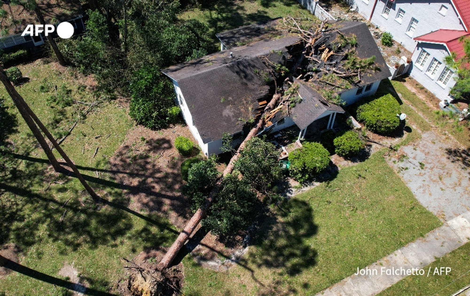 Aerial view of a residential home that has sustained significant damage from a powerful storm, with a large tree having fallen on its roof. The house is surrounded by a grassy yard, with patches of greenery and hedges visible. The tree, which lies diagonally across the roof, has caused considerable destruction, with branches and debris scattered around. Nearby, another house with a red roof can be seen to the right, adding context to the neighborhood setting. The image captures the aftermath of storm Helene, which has caused severe impacts in the area, including loss of life and extensive property damage.