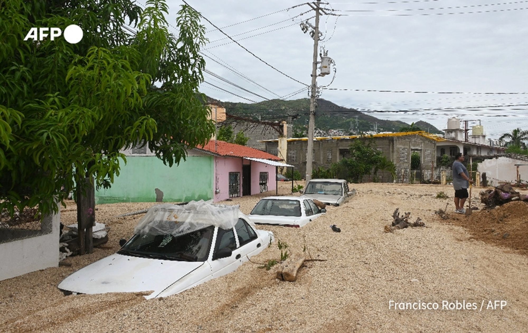 A scene depicting the aftermath of Hurricane John, showing significant flooding in a residential area. Several white cars are partially submerged in a muddy mixture, with only the roofs visible above the water level. The surrounding landscape is littered with debris, and a man can be seen in the foreground, standing and presumably inspecting the damage as he holds a shovel. In the background, there are two colorful houses, one painted pink and the other green, with a power pole and overhead electrical wires stretching above them. The sky is overcast, adding to the somber atmosphere of the scene, which reflects the devastating impact of the hurricane in the area.