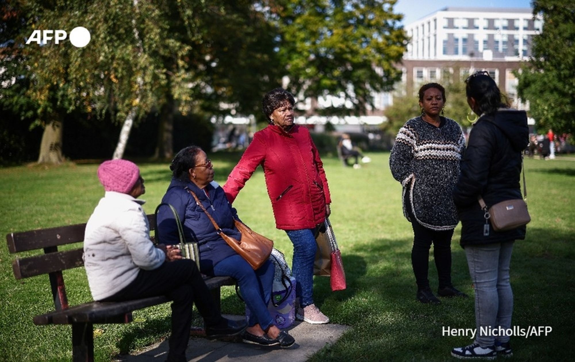 A group of four women, of African descent, is gathered in a sunny park setting. Two women are seated on a wooden bench, while two others stand nearby, engaged in conversation. 

The first seated woman on the left is wearing a white jacket, and has her arm resting on the back of the bench. Next to her, another woman is dressed in a navy coat and is holding a brown bag. The third woman, standing, is in a bright red coat with a matching handbag, and appears to be speaking animatedly. She has curly hair and is looking towards the fourth woman. 

The fourth woman, who also stands, is dressed in a black and white patterned sweater, black pants, and has her hair tied back. She appears to be listening attentively. The background features green grass, trees, and a modern building, suggesting a vibrant community space. The atmosphere is friendly and lively, indicative of a social gathering.