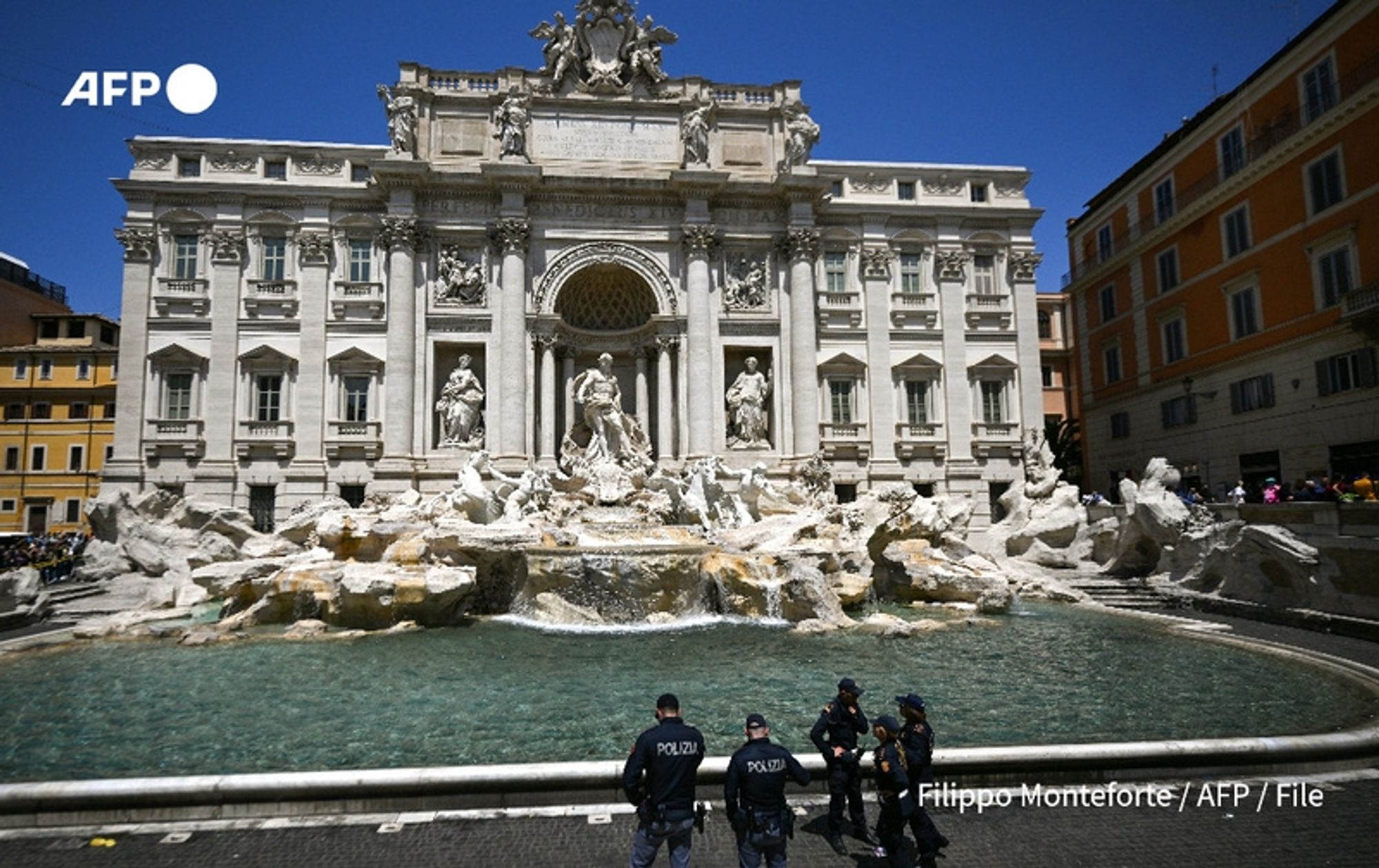 The image shows the magnificent Trevi Fountain, a Baroque masterpiece located in Rome. The fountain features an elaborate façade adorned with sculptures of mythical figures, including the central figure of Oceanus, who rides a chariot pulled by sea horses. The intricate stonework displays detailed carvings, enhancing the grandiosity of the fountain. The water cascades from various points, creating a tranquil ambiance.

In the foreground, a group of police officers, identifiable by their uniforms marked with "POLIZIA," are standing near the fountain, overseeing the area. The surrounding plaza is bustling with visitors admiring the beauty of the fountain, many engaging with its iconic tradition of throwing coins. The buildings flanking the fountain showcase classic Roman architecture, with tan, pale-yellow façades contrasting against the clear blue sky.

The scene captures a sunny day, highlighting the vibrant atmosphere of this historic location. The overall impression is one of splendor and cultural significance, reflecting the Trevi Fountain's status as a must-see landmark in Rome.