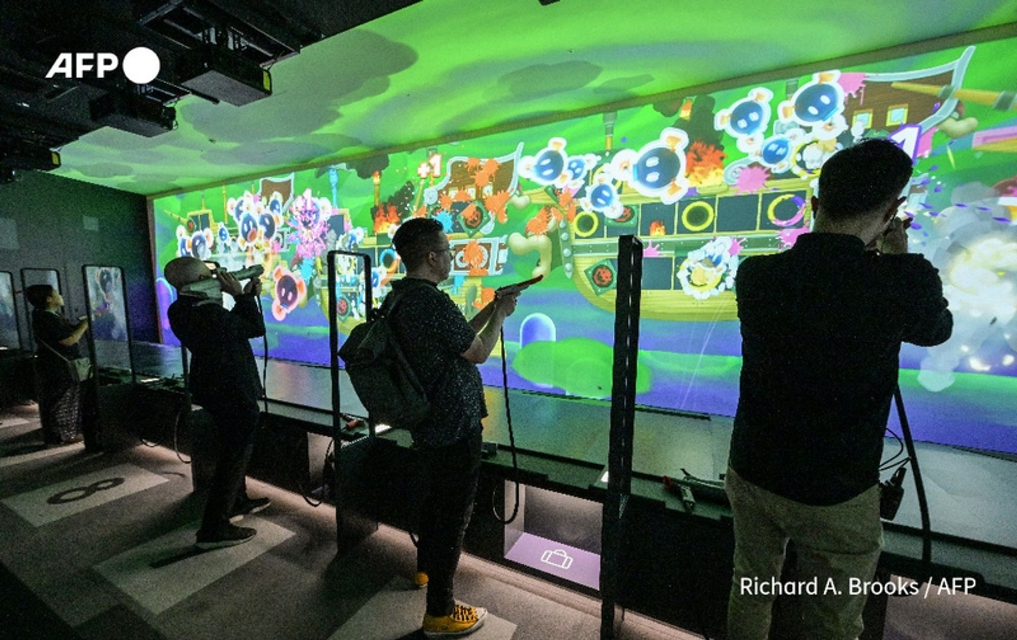 Three individuals are engaged with an interactive display at Nintendo's newly opened museum in Kyoto. The background features a vibrant, animated projection showcasing colorful characters and scenes inspired by Nintendo games, including elements resembling cheerful animals and fantastical landscapes. Two men are positioned close to the display: one on the left, wearing a black shirt and holding a device, and the other in the center, dressed in a patterned shirt and pointedly aiming a gaming peripheral towards the screen. A third person is situated towards the right, capturing the experience with a camera. The atmosphere is lively and immersive, reflecting the richness of Nintendo's gaming history and engagement with its audience. The interior of the museum is modern, with a sleek design and dim lighting that highlights the bright visuals on the wall.