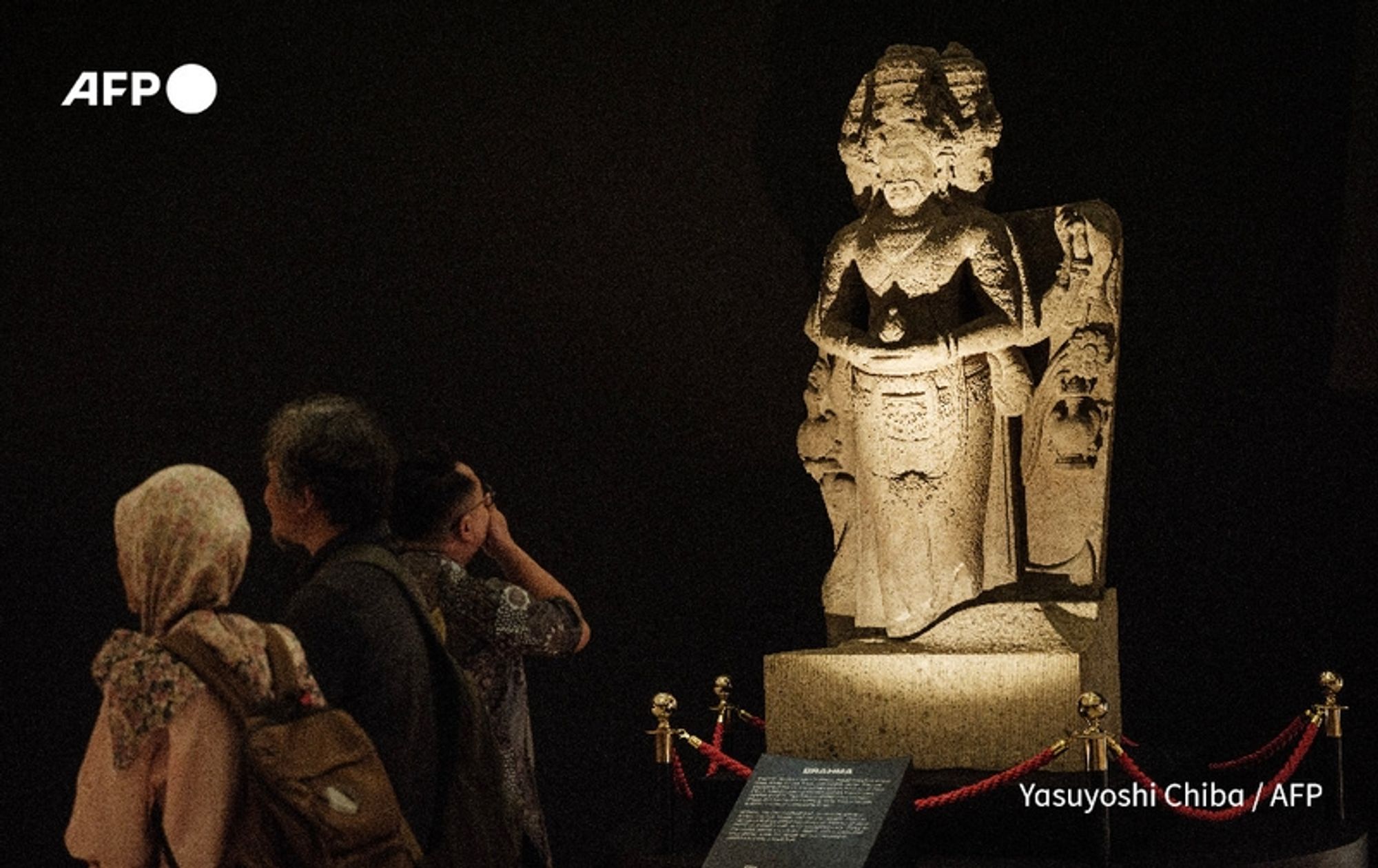 A dimly lit exhibit in the National Museum of Indonesia in Jakarta showcases a large, intricately carved statue of Brahma, the Hindu deity. The statue, made from stone, stands prominently in the center of the image, exhibiting detailed features, including multiple arms and ornate attire. Surrounding the statue are several visitors, some observing intently and taking photographs. The visitors include a woman wearing a patterned headscarf and a group of individuals dressed in casual clothing. A small plaque in front of the statue provides information, partially obscured by the exhibit's protective ropes. The overall atmosphere conveys a sense of reverence, highlighting the importance of the repatriated artifacts on display, amidst a backdrop of rich cultural history.