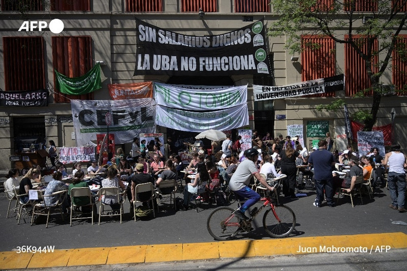 A group of Argentine university professors and students are participating in a street protest in Buenos Aires, advocating against President Javier Milei's veto of increased funding for public universities. The scene is vibrant, with various banners hanging from a building that includes slogans such as "Sin Salarios Dignos" (Without Dignified Salaries) and "La UBA No Funciona" (UBA Does Not Function). The banners are colorful and display messages supporting the need for better funding in education. 

In the foreground, a large gathering of people is seated at tables, engaged in discussions and activities related to their cause. The setting is lively, with individuals of diverse backgrounds, some taking notes, others interacting animatedly. To the right, a person is riding a bicycle, adding to the dynamic atmosphere of the protest. The sidewalk is lined with chairs and tables, while trees provide some shade. The overall mood conveys determination and a strong sense of community among those advocating for educational support.