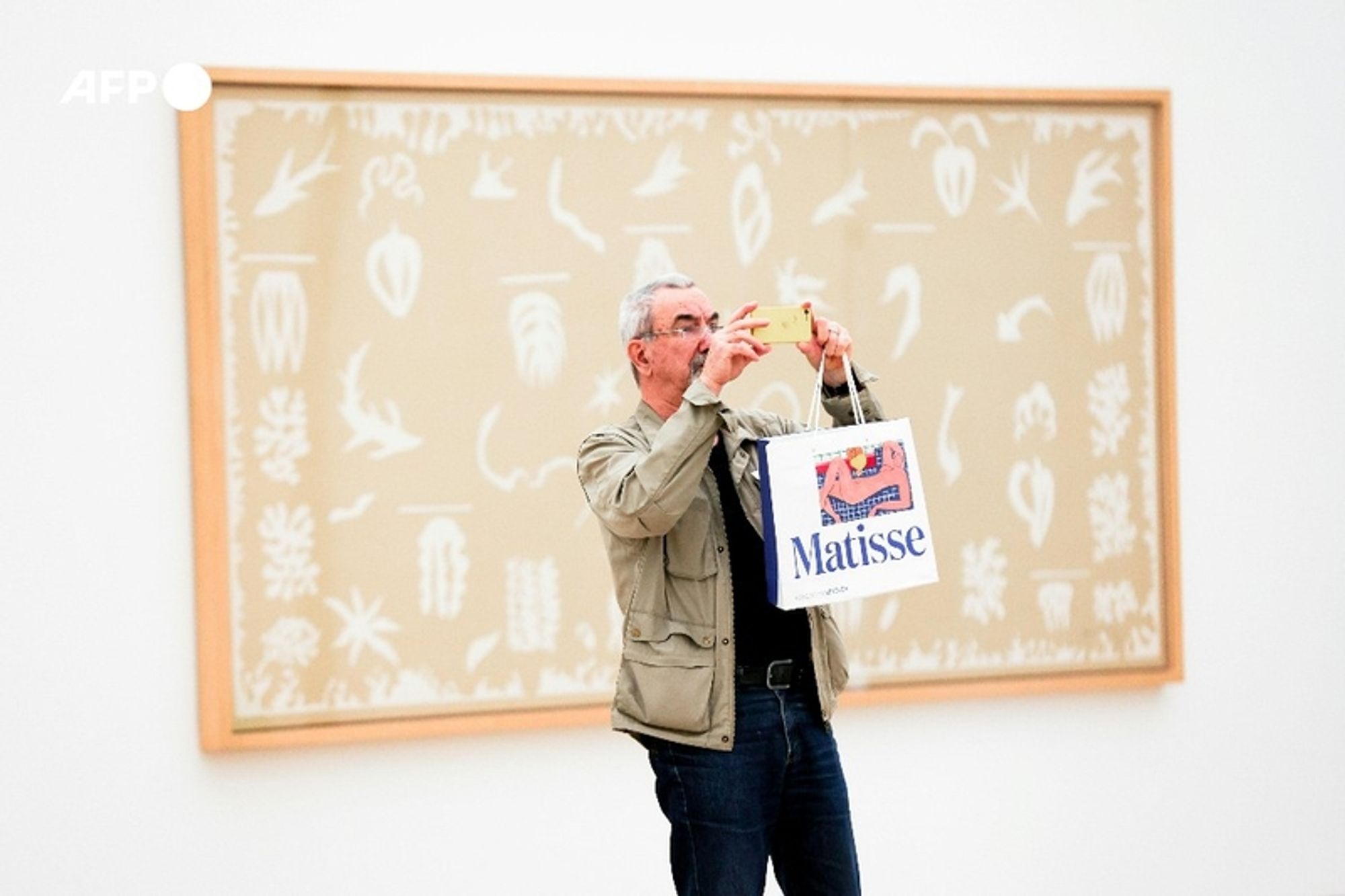 A middle-aged man with gray hair and glasses stands in a spacious art gallery, taking a photograph using his smartphone. He is dressed in a light gray jacket, a black shirt, and blue jeans. In his left hand, he holds a white shopping bag with a colorful graphic and the name "Matisse" prominently displayed on it. Behind him is a large artwork by Henri Matisse, featuring a beige background adorned with delicate white line drawings of various botanical and abstract shapes. The frame of the artwork is a simple wooden design that complements the piece's aesthetic. The gallery has neutral-colored walls, enhancing the focus on the art displayed. The environment appears bright and well-lit, reflecting a contemporary artistic space.