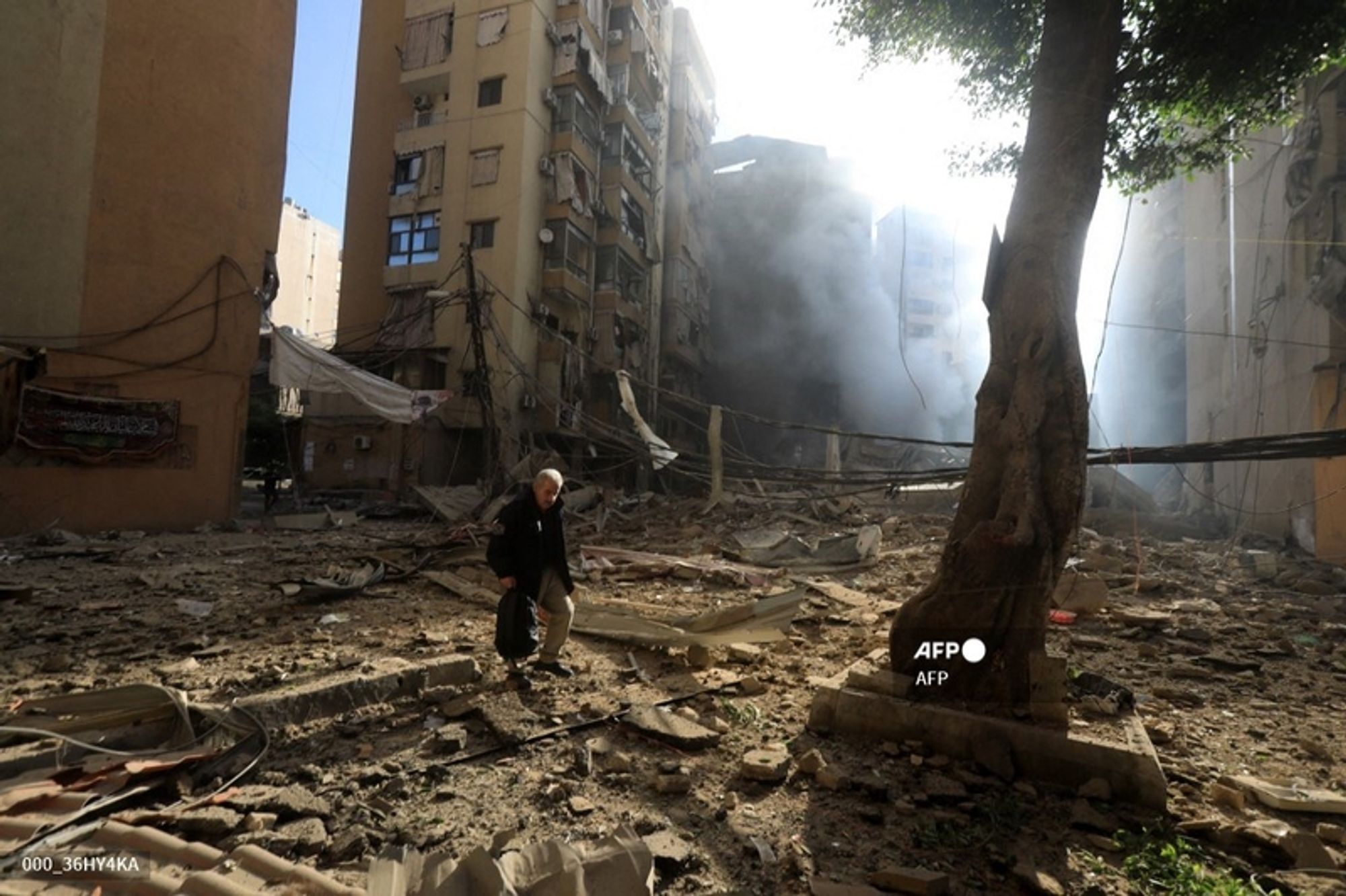 A man with gray hair walks through a devastated urban landscape, carrying a black bag. The scene is marked by considerable destruction, with debris scattered across the ground and parts of buildings in ruins. Tall residential buildings, whose walls are scarred and shattered, rise in the background, with some windows broken and debris hanging from the edges. Dust and smoke billow from the wreckage, suggesting a recent explosion or disaster. A tree stands nearby, providing an element of contrast against the destruction. The stark sunlight streams into the scene, illuminating the dust and providing a surreal ambiance to the grim reality of the environment. The overall atmosphere is somber, highlighting the severity of the situation depicted in the image.