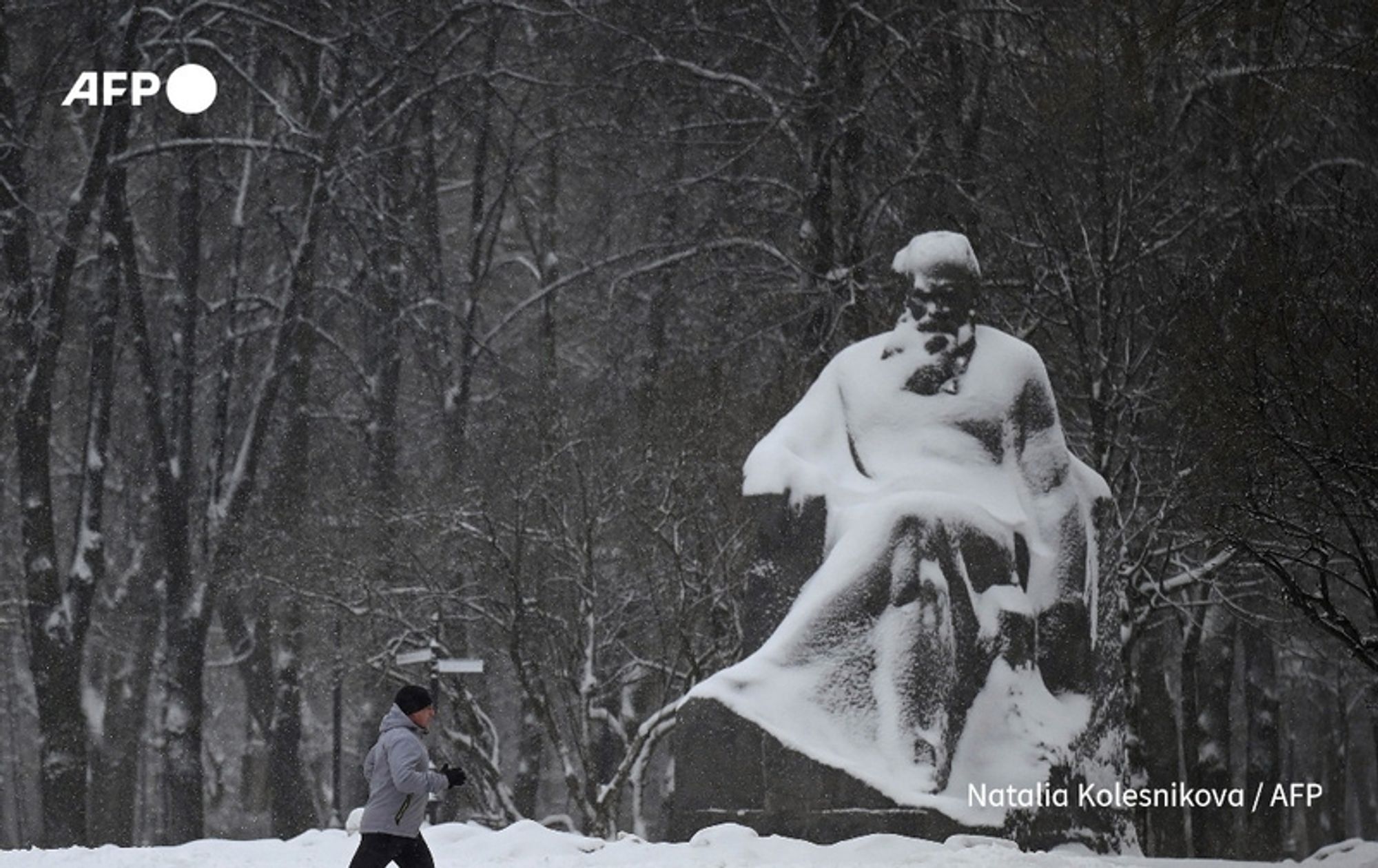 A snowy scene depicting a solitary figure walking in a winter landscape. The person is dressed in a gray hooded jacket and black pants, briskly walking past a large statue partially covered in snow. The statue, resembling the famous Russian author Leo Tolstoy, depicts him seated with a contemplative expression, wrapped in his clothing. Surrounding the statue are tall, bare trees, and light snowfall can be seen falling gently in the background, creating a quiet and serene atmosphere. The image captures the somber beauty of winter while evoking the legacy of Tolstoy amidst a dialogue about peace in present-day Russia.