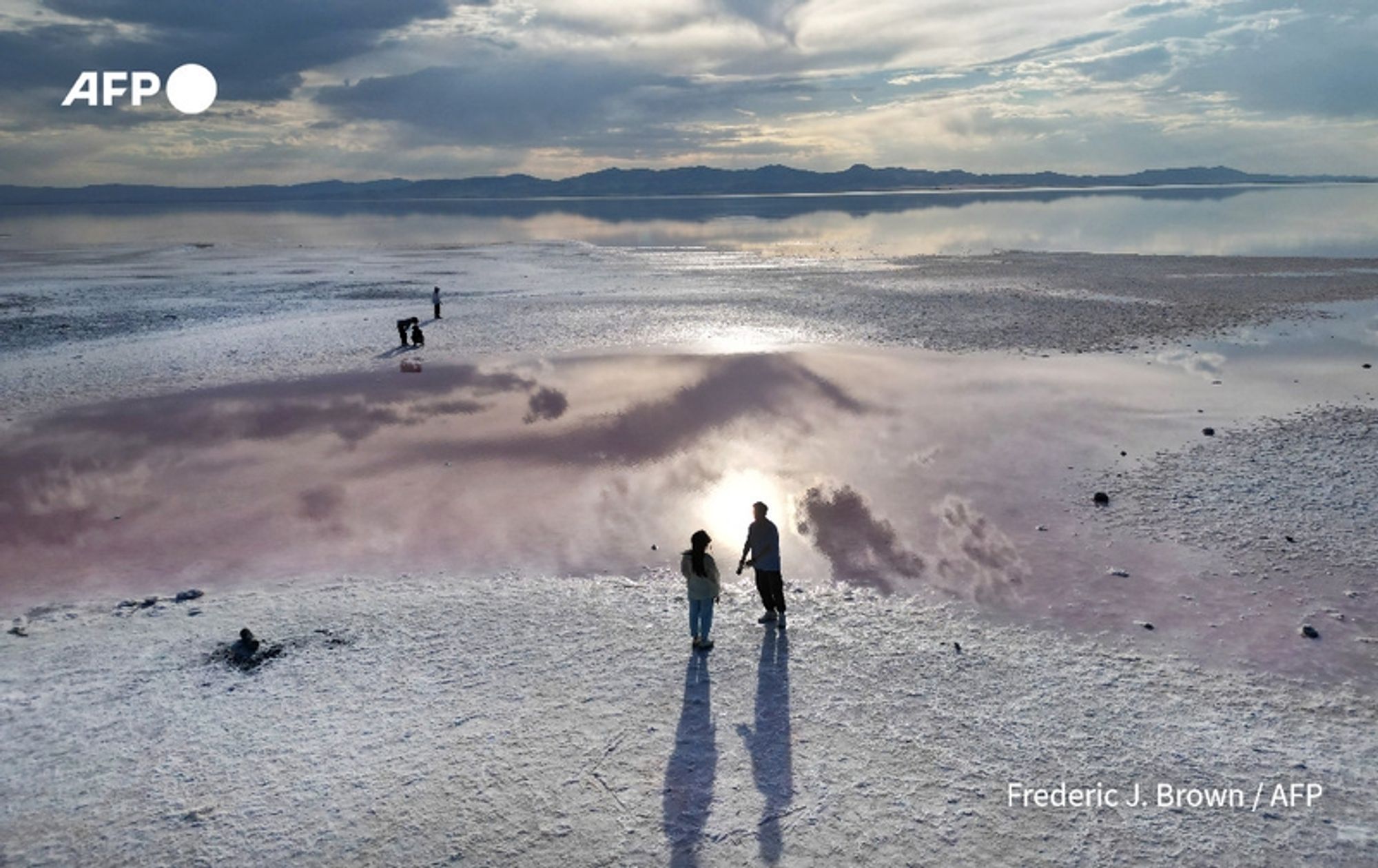 A wide aerial view of the Great Salt Lake in Utah, partially dried, showcasing its unique landscape. In the foreground, two silhouetted figures stand on the cracked, white salt flats, looking out over the lake’s pinkish waters which reflect the clouds above. Their long shadows stretch out behind them on the salt surface. A third person can be seen in the distance, near the shoreline, capturing the scene with a camera. The background features rolling mountains and a dramatic sky filled with scattered clouds, highlighting the area’s natural beauty amidst the environmental changes.