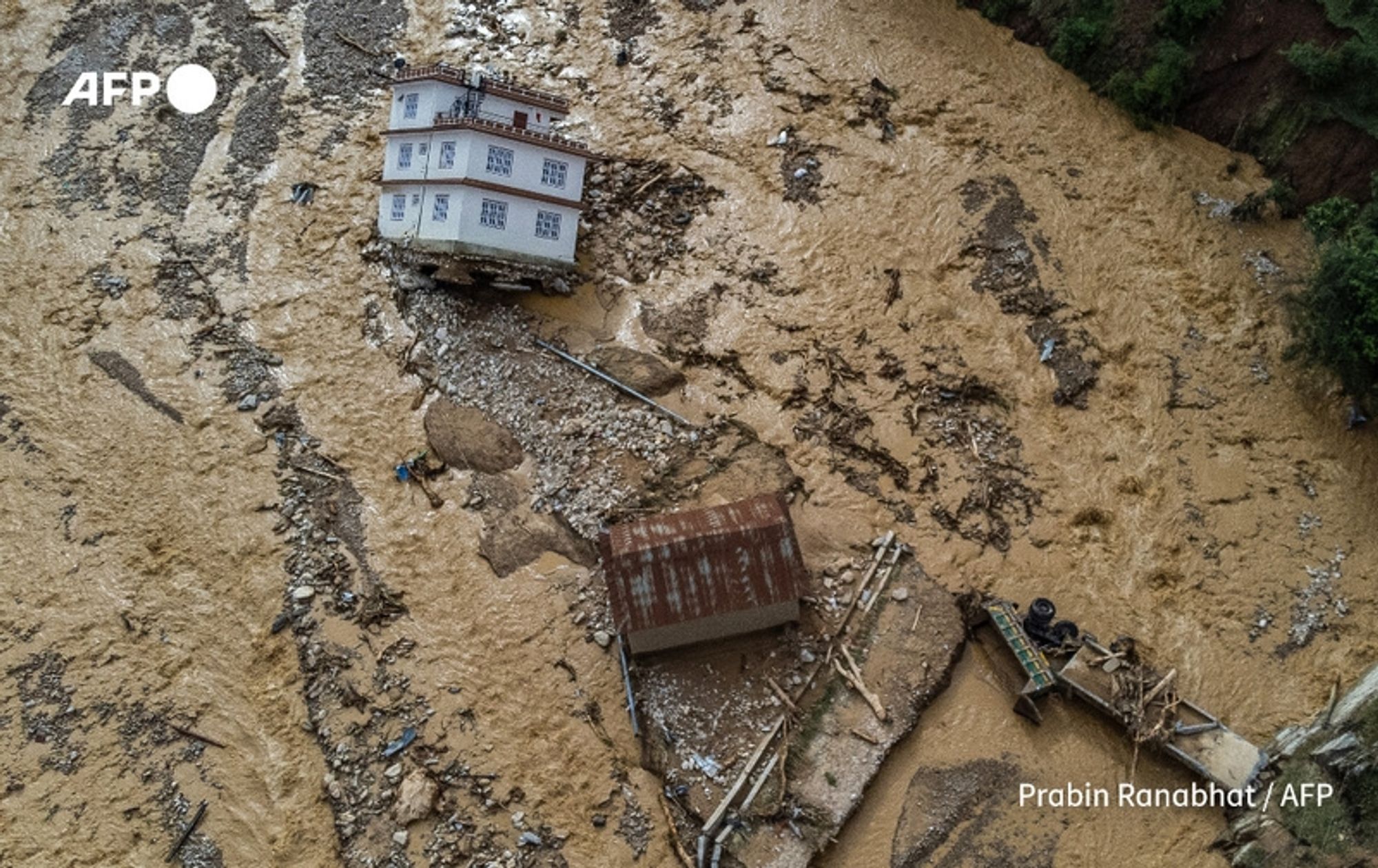 Aerial view of a severely affected area in Nepal after monsoon floods. The image shows two houses amidst a landscape flooded with brown, muddy water. One house, situated on a small rise, appears to be partially submerged and is tilted, suggesting it has been destabilized by the flood. The second house, smaller and with a rust-colored roof, is surrounded by debris, including broken timber and other materials washed away by the floods. The surrounding terrain is strewn with mud and stones, highlighting the extent of the destruction. The cloudy sky looms above, adding to the somber atmosphere of the scene. In the distance, patches of greenery are visible, contrasting with the devastation below.