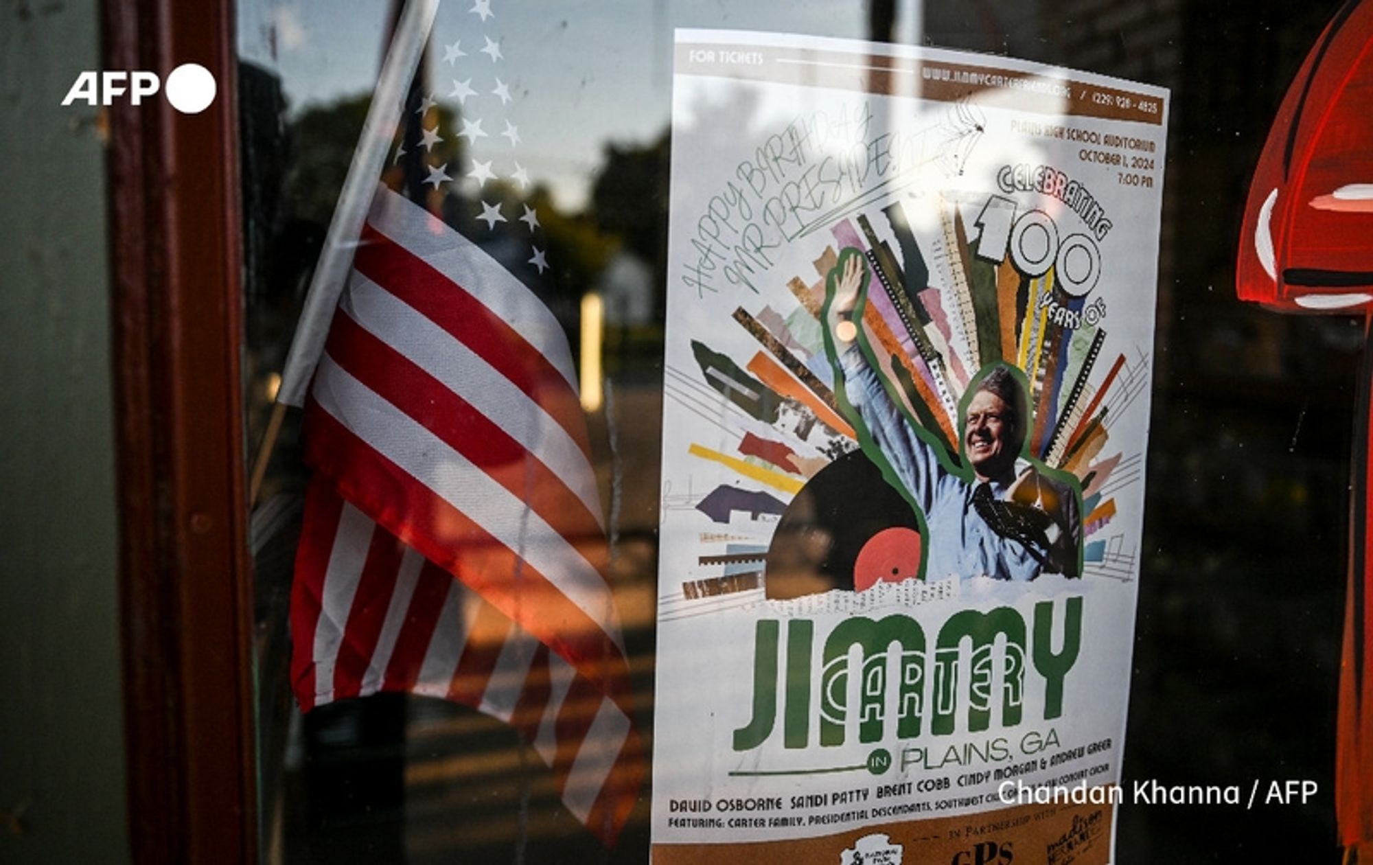 A close-up view of a reflective window displaying a colorful poster celebrating Jimmy Carter’s 100th birthday. The poster features a large image of Carter smiling and waving, with vibrant rays of color emanating from behind him, symbolizing joy and celebration. Above him, the text reads "Celebrating 100 Years," and below, "JIMMY CARTER" is prominently displayed in large, bold green letters, followed by "PLAIN, GA." Handwritten notes in black pen express birthday wishes, saying, "Happy Birthday Mr. President!" The background includes an American flag draped in the window, its red, white, and blue colors visible. This scene is set in Plains, Georgia, indicating the hometown pride for its most famous resident, as the community prepares for a significant celebration.