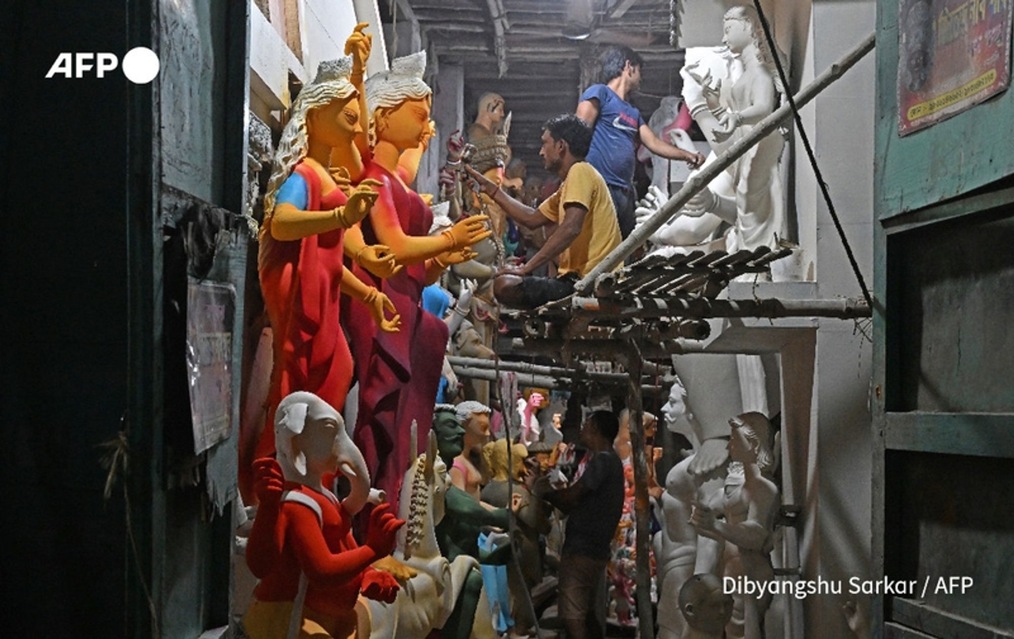 Amidst a bustling workshop in Kolkata, colorful statues of Hindu deities and figures sit prominently displayed. The scene shows artisans at work, with a man perched on a scaffold, carefully painting a statue. The vibrant colors of the figures contrast with the more muted tones of the workshop's surroundings. Statues of various sizes, including some with intricate detailing, can be seen lining the walls, showcasing traditional craftsmanship. The lighting is dim but highlights the vivid hues of the statues, creating a sense of vibrancy and life within the space. Various tools and paintbrushes are scattered around, reinforcing the atmosphere of artistic creation. The environment reflects a blend of cultural reverence and the labor involved in preparing these figures for the upcoming Hindu rituals, emphasizing the intersection of tradition and the daily lives of the artisans.