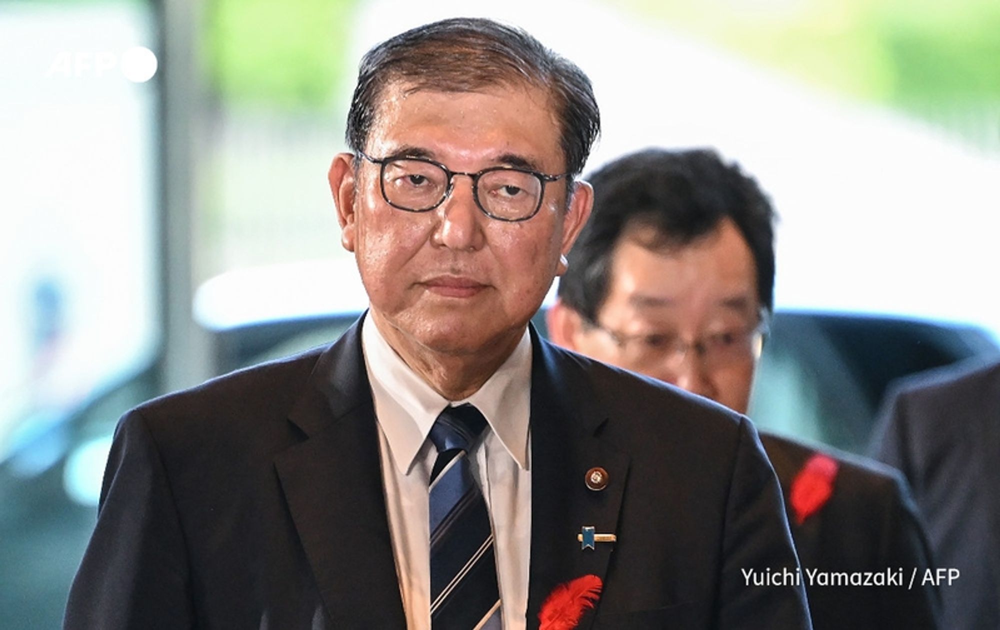 A man with short, slicked-back hair and glasses stands prominently in the foreground, wearing a dark suit with a white dress shirt and a dark striped tie. He has a red flower pinned to the left side of his suit jacket, suggesting a formal occasion or event. His expression is serious and contemplative as he gazes directly at the camera. In the background, a blurred figure can be seen, also dressed in formal attire, indicating a gathering or event involving officials. The setting appears to be well-lit, with natural light filtering in, suggesting an indoor environment. The image conveys a sense of importance, likely related to political affairs in Japan.