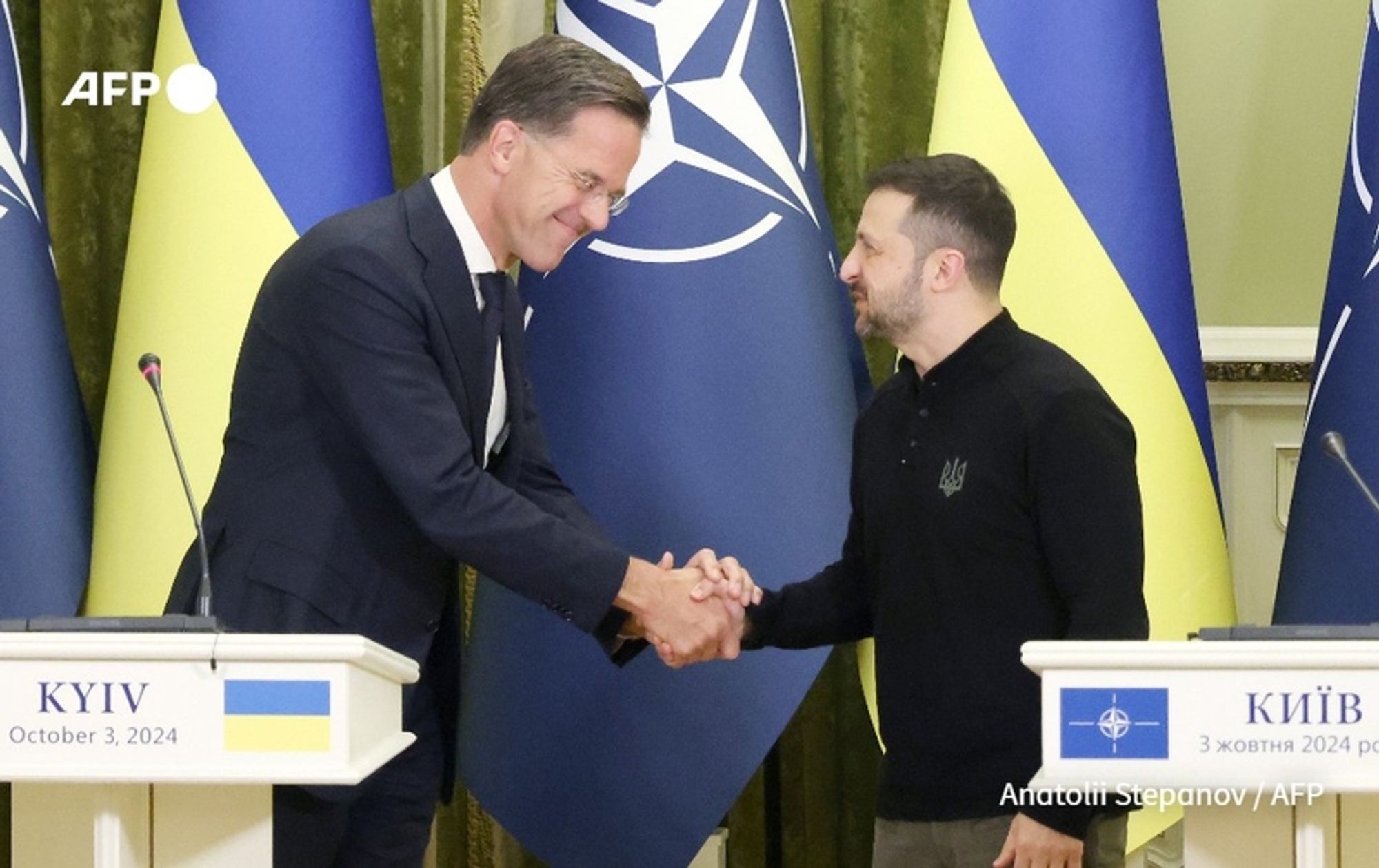 A diplomatic meeting between Mark Rutte, the Prime Minister of the Netherlands, and Volodymyr Zelensky, the President of Ukraine, takes place in Kyiv on October 3, 2024. Both leaders are engaging in a handshake, with Rutte on the left, dressed in a dark suit and tie, and Zelensky on the right, wearing a casual black sweater. The background features the Ukrainian flag in the form of vertical stripes, alternating blue and yellow, and a large NATO emblem prominently displayed. The scene gives an impression of collaboration and solidarity, as both leaders smile and appear engaged in a positive exchange. A podium with a white tablecloth is positioned between them, but the specific details of the text on the podium are not visible. The atmosphere reflects a moment of unity and support for Ukraine amidst ongoing challenges.