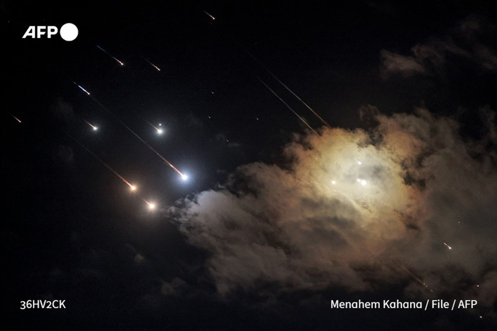 A dark night sky filled with streaks of light representing missiles or projectiles being launched. The image captures multiple bright points moving diagonally from the upper left toward the lower right, suggesting a rapid upwards trajectory. A grayish cloud is visible in the center, illuminated by flashes that could indicate explosions or impacts. The surrounding atmosphere appears somber, with a mix of dark clouds and bright lights contrasting against the night. The image is attributed to Menahem Kahana, with the logo "AFP" positioned in the upper left corner.