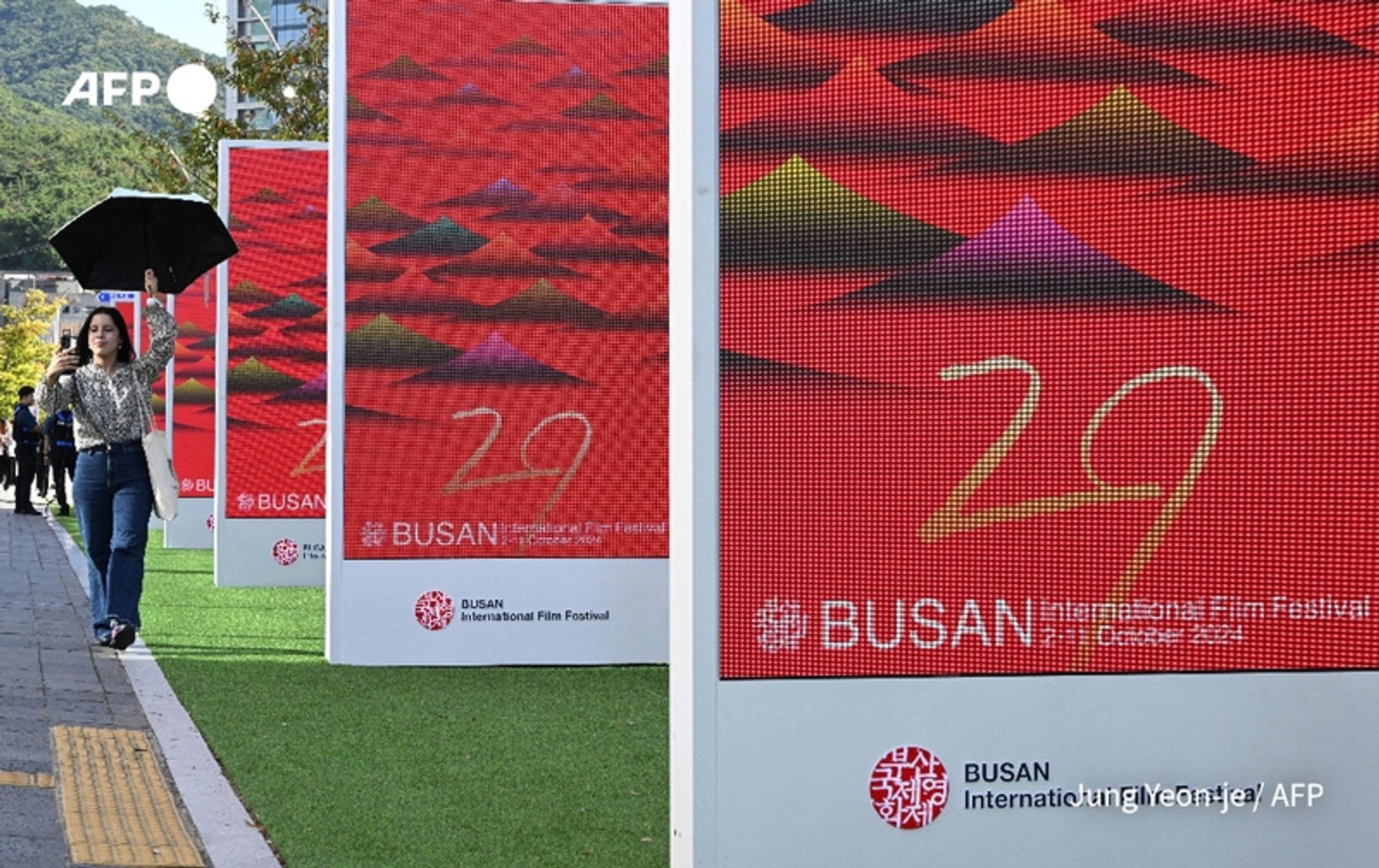 A woman holding a black umbrella walks along a pathway at the Busan International Film Festival venue. She is dressed in casual attire and is seen passing by large promotional screens. The screens display colorful graphics, prominently featuring the number "29" in a playful, cursive font, alongside the words "BUSAN International Film Festival." The background colors of the screens include vibrant reds, greens, and purples, depicting stylized mountain shapes, adding an artistic touch. The scene is set outdoors with a backdrop of lush, green hills under clear skies. The area is lined with grass, contributing to a festive atmosphere. The event, which is significant as it marks the opening of Asia's largest film festival, emphasizes a blend of cinema and culture.