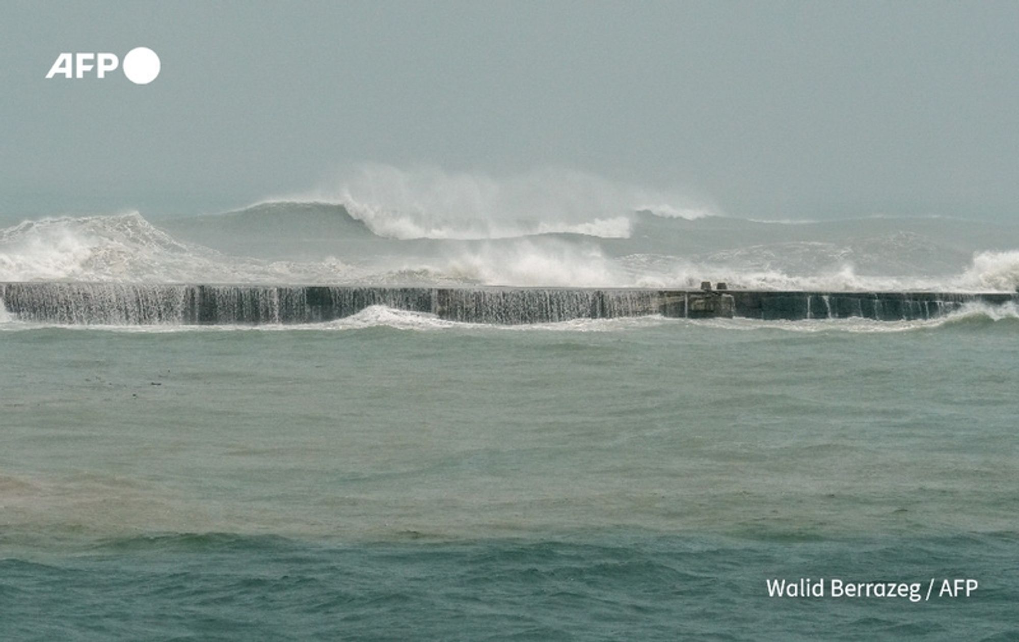 A turbulent seascape depicting large, crashing waves battering a coastal breakwater. The foreground shows the dark, tumultuous water with white foam cascading off the top of the breakwater, illustrating the powerful impact of the waves. The sky is overcast with a greyish hue, contributing to a sense of impending storminess. The scene conveys the intensity of Typhoon Krathon as it approaches Taiwan, emphasizing the rough seas and adverse weather conditions. The backdrop suggests a challenging maritime environment, indicative of the typhoon's effects on the region. In the corner of the image, a logo reading "AFP" is visible.