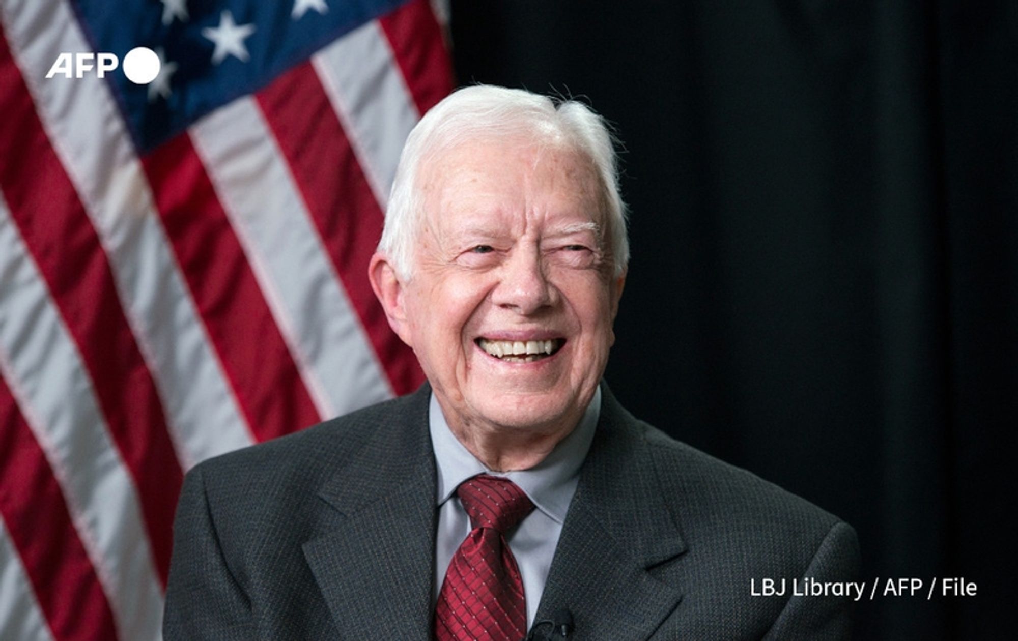 A smiling elderly man with white hair is seated in front of a dark backdrop, accompanied by a large American flag to his left. He is wearing a dark gray suit with a light blue shirt and a maroon tie. The man appears warm and approachable, with a broad smile that highlights his cheerful demeanor. His expression conveys a sense of joy and contentment, reflecting the celebratory context of his 100th birthday. The American flag behind him features red and white stripes and blue with white stars, symbolizing his status as a former U.S. president.