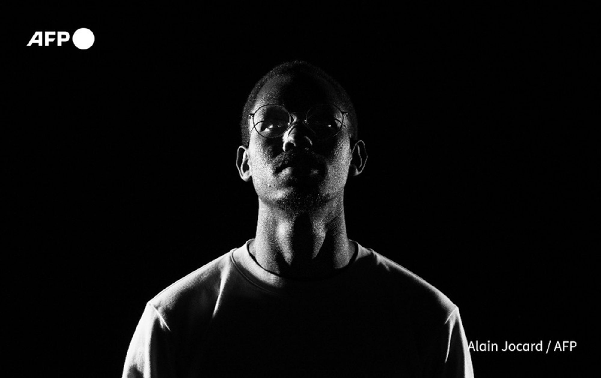 A black and white portrait of a man, Nelson Amenya, who is the whistleblower regarding secret negotiations for an Indian company to take over Kenya's main airport. He stands against a solid black background, with only his silhouette illuminated from below, highlighting his facial features and glasses. His expression conveys a sense of seriousness and contemplation, reflecting the gravity of his situation. He wears a round-necked shirt, and his body is slightly turned to the side. The dramatic lighting creates a stark contrast, emphasizing the tension and fear he faces following his revelations. In the top left corner, there is a logo "AFP" in white.