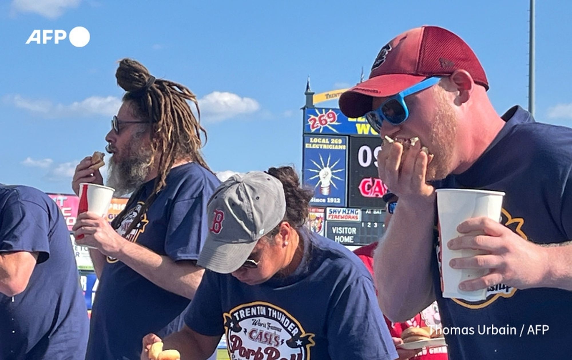 Three participants are shown engaged in a competitive eating contest featuring pork rolls. The setting is outdoors, likely at a fair or festival, under a clear blue sky. 

On the left, a man with long, dreadlocked hair wears sunglasses and a dark t-shirt. He holds a pork roll close to his mouth, focused on eating. 

In the center, a woman with dark hair pulled back into a ponytail sports a light gray baseball cap and a navy t-shirt. She appears to be taking a bite of her pork roll while holding a cup in her other hand. 

On the right, a man with a short beard wears a maroon cap and sunglasses. He is also eating a pork roll and has his hand near his mouth, possibly indicating he is in the midst of chewing. 

In the background, there are colorful banners and signs, likely indicating the event's details, with a large score sign visible overhead. The scene captures the excitement and competitive spirit of Major League Eating, as the competitors focus on consuming as many pork rolls as possible within the time limit.