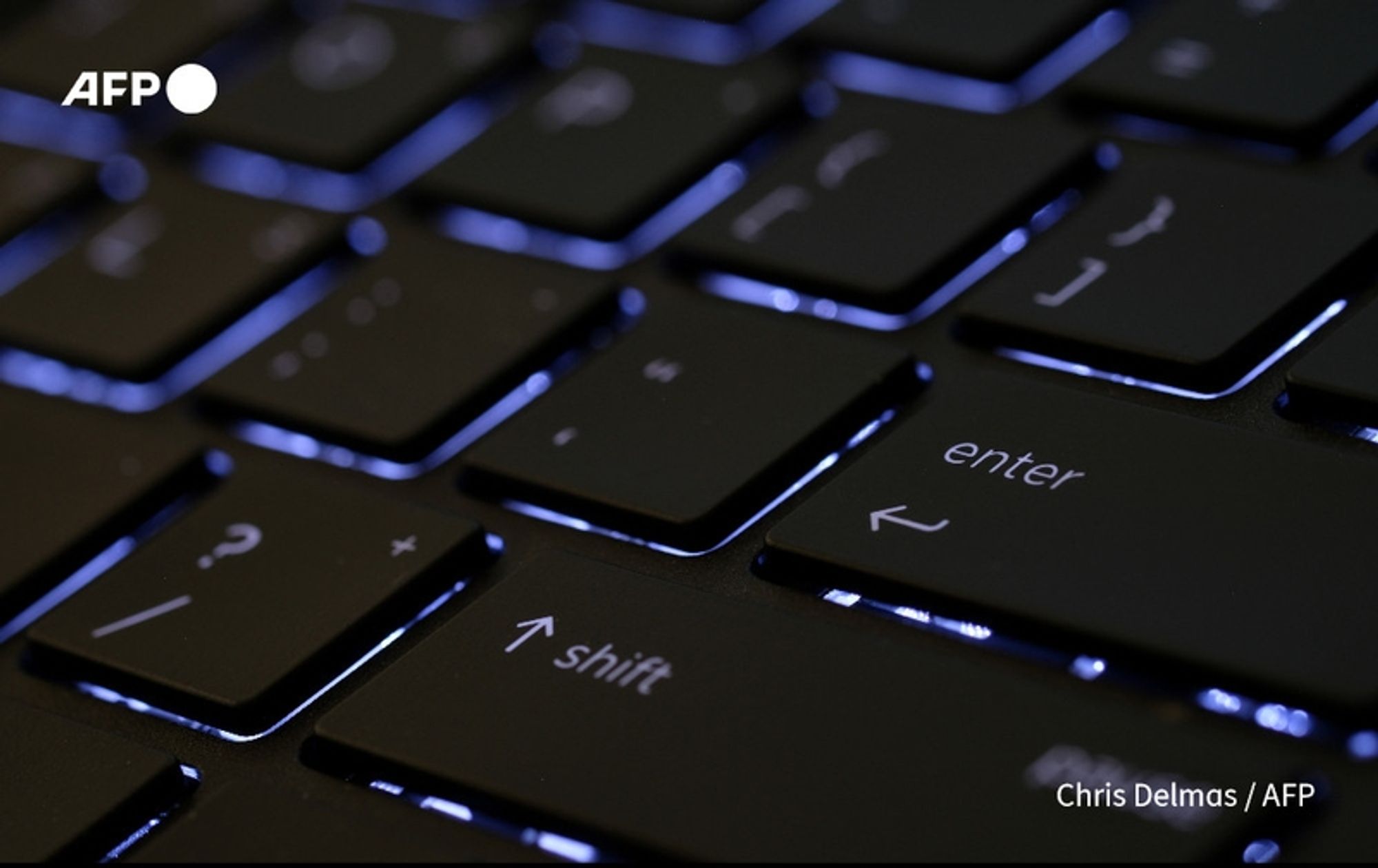 A close-up image of a black computer keyboard, showcasing several illuminated keys. The keys predominantly exhibit a shiny, dark surface with faint reflections of light, highlighting their contours. The "enter" key is prominently visible, located in the center of the image, along with the "shift" key to the left. The keys feature white symbols and letters, providing contrast against the dark background. The background is blurred, emphasizing the keyboard. In the top left corner, the logo "AFP" is displayed. The overall tone of the image conveys a focus on technology and cybersecurity, relevant to the context of a cyberattack on the Internet Archive.