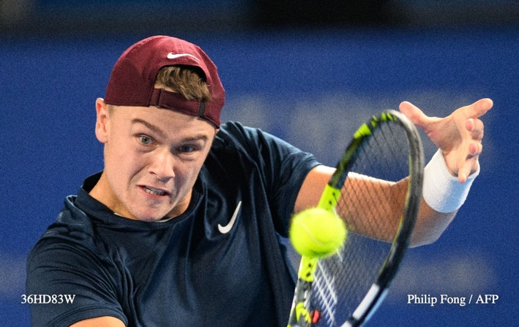 A young male tennis player, Holger Rune, is captured mid-action on a bright tennis court. He is wearing a dark blue athletic shirt with a white logo and a maroon cap. His facial expression is focused and intense as he prepares to strike a yellow tennis ball, which is positioned just in front of him. His left arm is extended forward, gripping the handle of a tennis racket, while his right arm is raised, ready to make contact with the ball. The background features a blurred audience and blue court markings, emphasizing the competitive atmosphere of the match. The image conveys a sense of determination and athleticism as Rune competes in a high-stakes moment during the Japan Open.