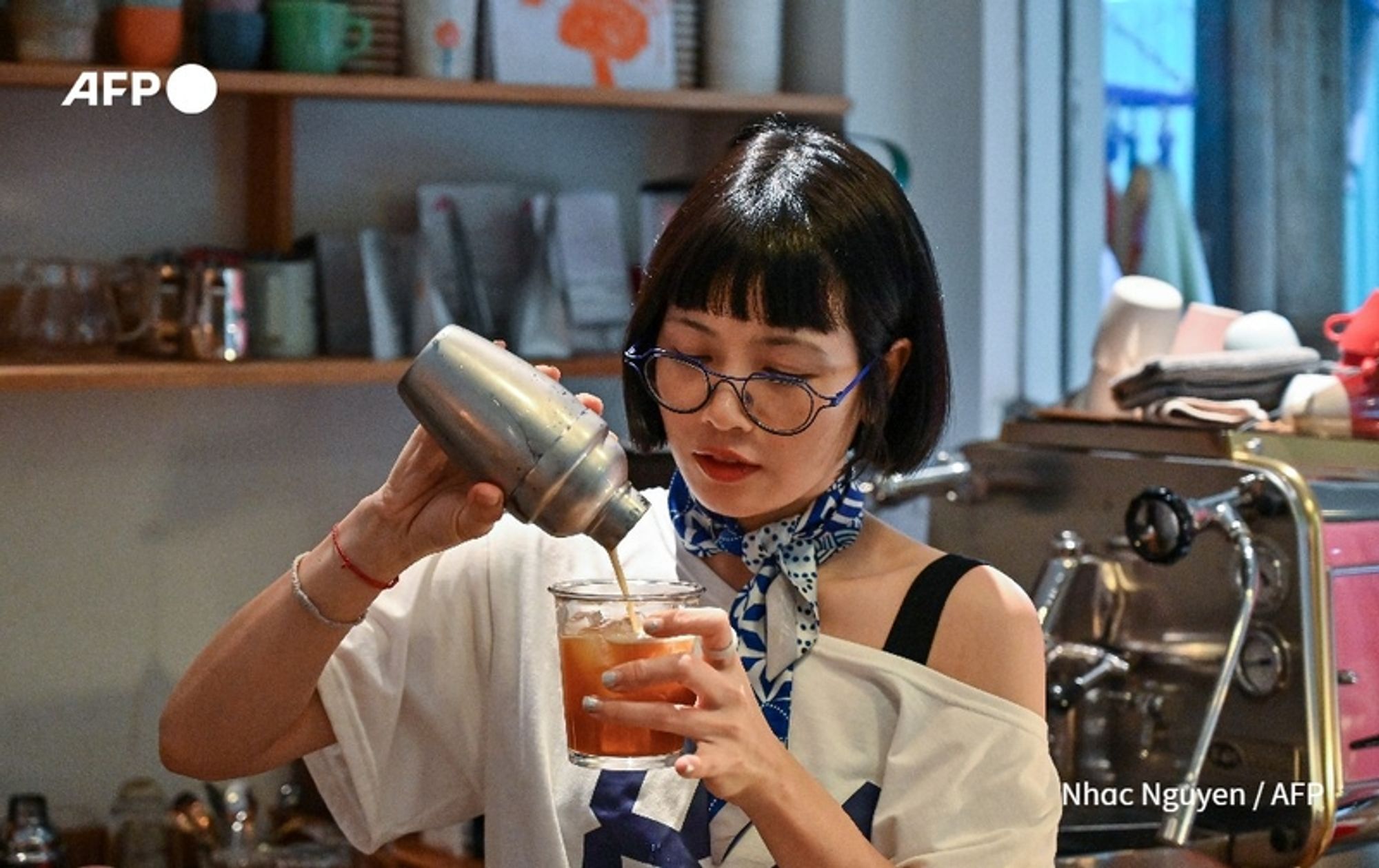 A young Vietnamese woman with short, straight black hair is expertly preparing a coffee drink in a café. She is wearing a loose-fitting white t-shirt with one shoulder exposed and has a blue bandana tied around her neck. Her round glasses enhance her focused expression as she pours from a shiny metal cocktail shaker into a clear glass filled with ice. The café has a modern and cozy atmosphere, with shelves in the background displaying various coffee-related items and decor. Natural light filters in from a window next to her, illuminating her work area and creating a warm, inviting ambiance.