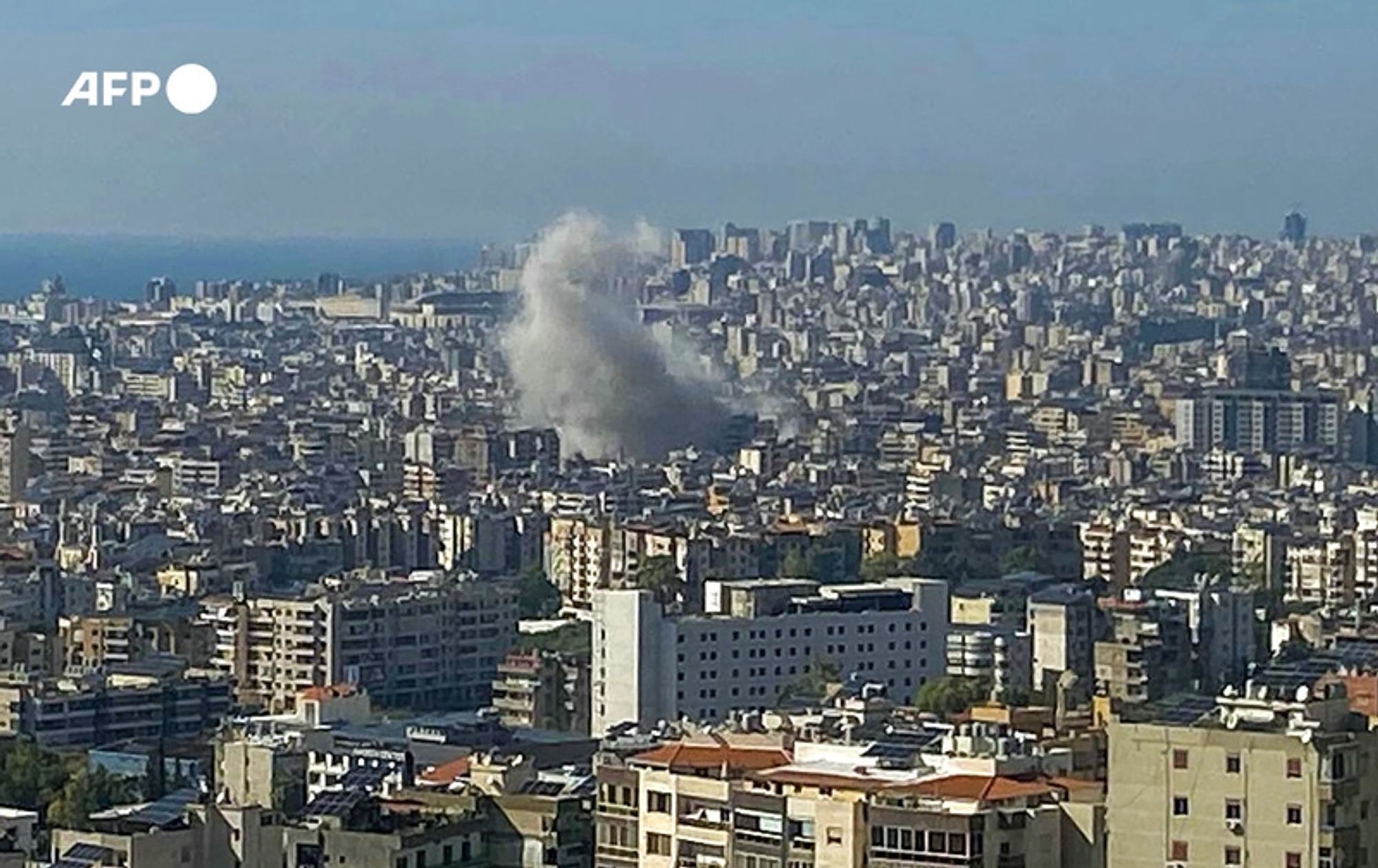 A panoramic view of a densely populated urban area, featuring numerous high-rise buildings and apartments clustered together, with a clear blue sky overhead. In the background, the deep blue waters of the sea are visible. The central focus of the image is a plume of smoke rising from a building within the city, indicating a recent explosion or military action. The smoke is grayish-white and billows upward, contrasting against the urban backdrop. The scene captures a moment of tension amidst the busy urban landscape, highlighting the impact of conflict in the area. The image is marked with a logo, "AFP," in the upper left corner.