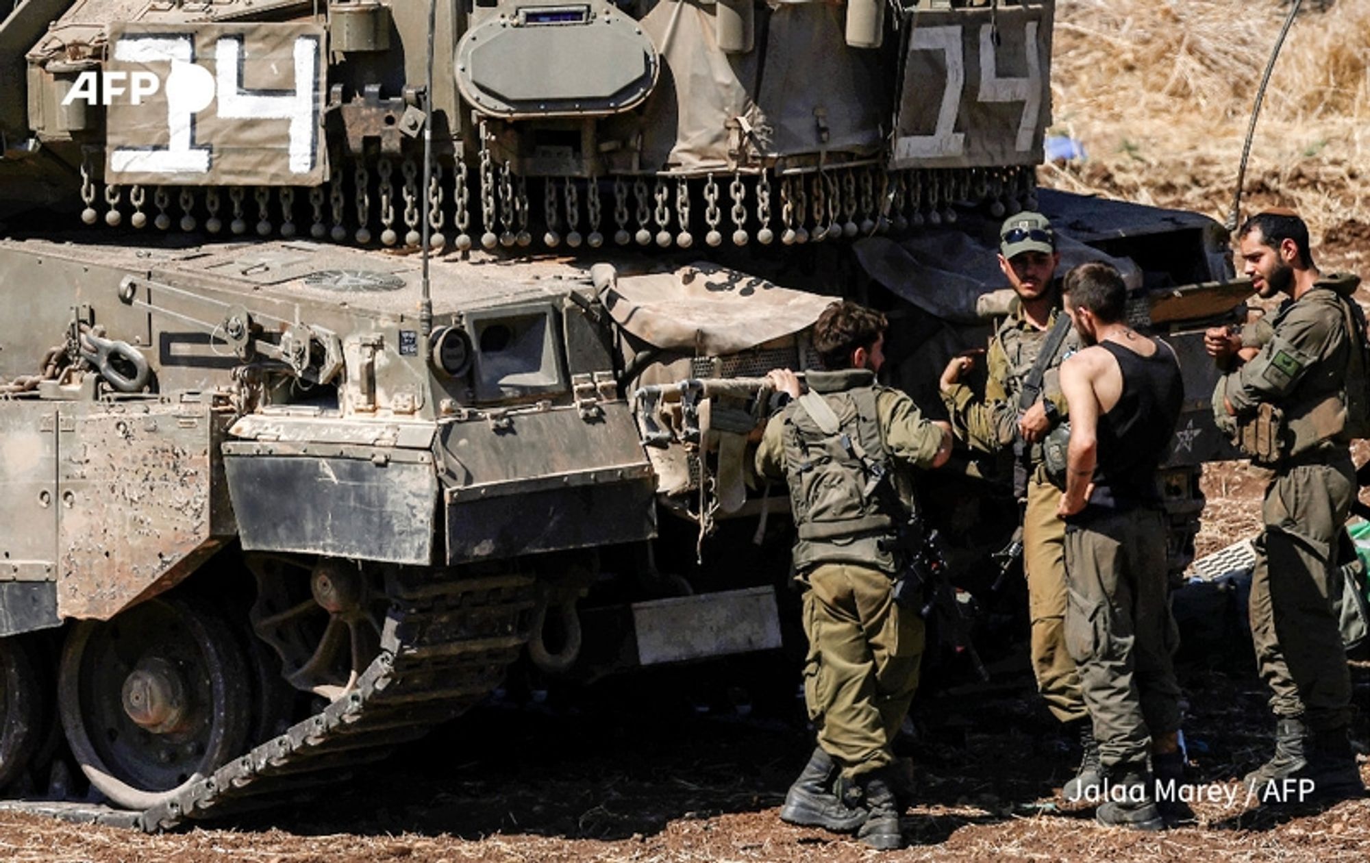 Four Israeli soldiers are gathered near a military tank in a dry, grassy area. The tank is heavily armored, with the number "24" prominently displayed on its side in white paint. The soldiers are in military uniforms; two are wearing caps, while one has rolled up sleeves and is holding a weapon. They appear to be engaged in conversation, with some focusing on the tank and others on each other. The setting suggests a tense environment, likely connected to ongoing military operations. The background is blurred, emphasizing the soldiers and the tank as the central elements of the scene.