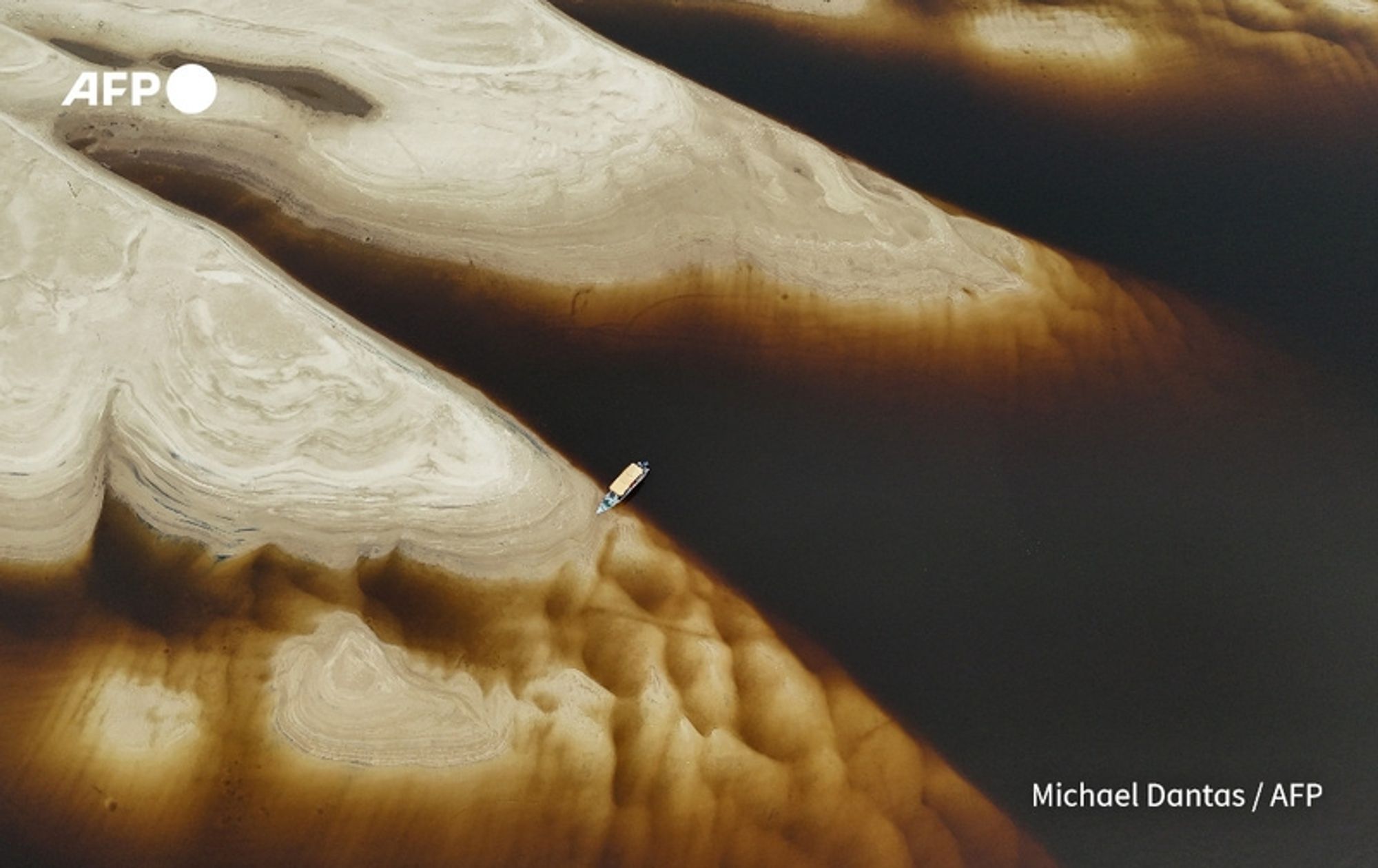 Aerial view of a dry riverbed on the Negro River in the Anavilhanas Archipelago, Amazonas state, northern Brazil. The sandy banks are displayed prominently, revealing various shades of beige and brown textures creating waves and grooves in the exposed land. In the foreground, a small boat is resting on a sandbank, its hull painted in light blue with a wooden deck. The contrasting dark water surrounds the sandy areas, reflecting deeper shades of brown. The landscape highlights the effects of a historic drought impacting several tributaries of the Amazon River, as noted by authorities. The image captures the stark beauty of the river's altered state in a visually striking manner. The upper left corner features the AFP logo.