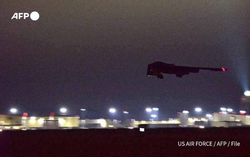 A B-2 stealth bomber is shown flying low against a night sky, with its dark silhouette prominently displayed. The background features a military airbase illuminated by numerous lights, highlighting various structures and operations within the facility. The scene conveys a sense of military activity and readiness. The image is marked with the logo "AFP" in the top left corner and includes "US AIR FORCE / AFP / File" in the lower right corner, indicating the source of the image.