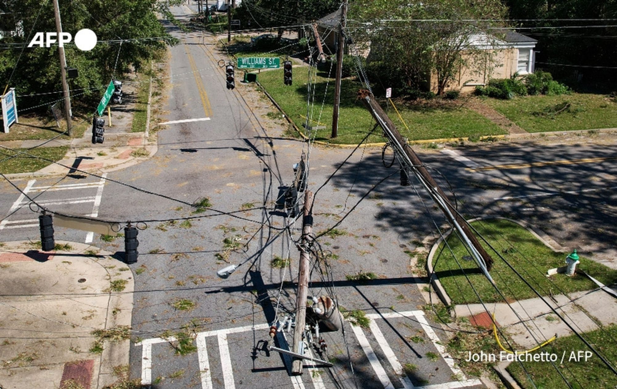 Aerial view of a street intersection in a neighborhood following the destructive impact of storm Helene. A utility worker is seen climbing a damaged utility pole, surrounded by downed power lines and debris scattered across the street. The road is lined with traffic lights, some non-functional, and a street sign indicates "WILLIAMS ST." In the background, several trees and buildings are partially visible, with branches down and visible damage from the storm. The scene reflects the aftermath of the storm, with signs of recovery efforts amidst the extensive disruption caused by the weather event.