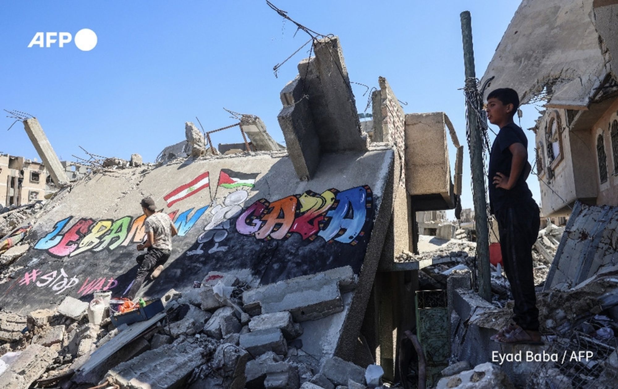 A scene depicting the aftermath of destruction in a war-torn area, where two young boys are observed amidst rubble and debris. The foreground features a large, fragmented wall, showcasing colorful graffiti that reads "LEBANON" and "GAZA," with a hashtag "#StopWar" beneath. The vibrant colors contrast starkly with the gray concrete debris surrounding them. One boy, dressed in a gray shirt and shorts, is climbing atop a mound of rubble, surveying the scene, while another boy, in a black shirt, stands confidently with his hands on his hips nearby. The background reveals a partially collapsed building, with exposed rebar and a clear blue sky above, emphasizing the destruction left behind by conflict.