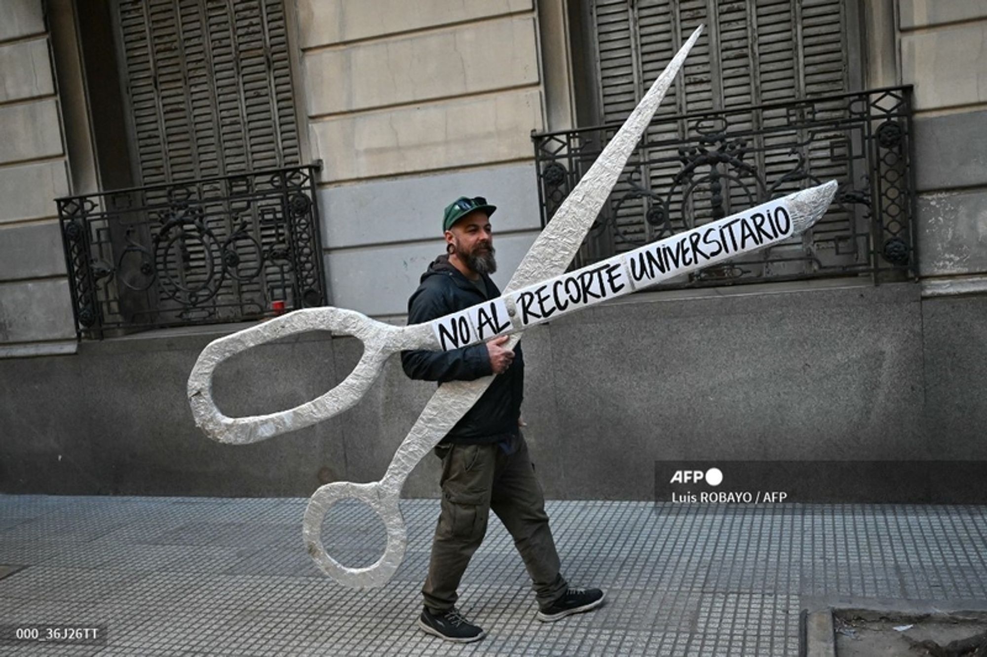 A man with a beard, wearing a black hoodie and a green cap, walks through a city street carrying a large, theatrical prop resembling oversized scissors. The scissors are painted white and feature a banner that reads "NO AL RECORTE UNIVERSITARIO," which translates to "NO to University Cuts," in bold black letters. The man is focused on the camera, walking past a building with decorative ironwork and tall windows in the background. The setting appears to be urban, emphasizing a public protest or demonstration regarding university funding issues. The ground is made of light-colored tiles, and the scene is well-lit, conveying an active atmosphere.