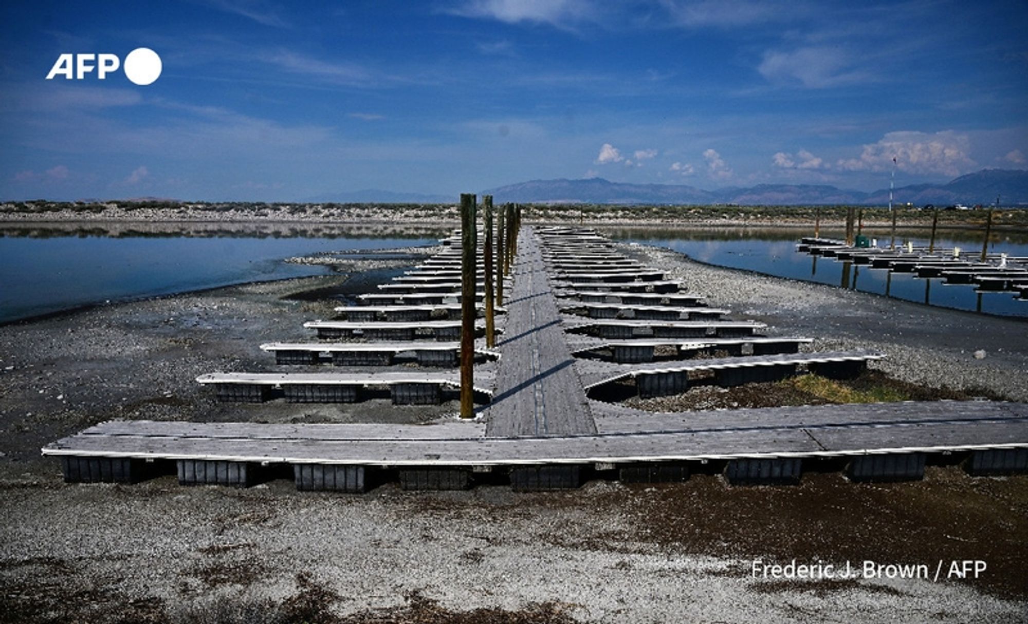 A wide-angle view of a partially dry dock area at the Great Salt Lake in Utah. The scene features a series of wooden boat docks that extend into shallow water, some sections appearing almost completely dry. The docks are arranged in a linear pattern, with several posts rising vertically, suggesting previous water levels. In the background, the expansive lake is visible, bordered by a distant mountain range under a clear blue sky with scattered clouds. The shoreline shows patches of dry earth and gravel, highlighting the lake's receding waters. The overall setting emphasizes the environmental changes impacting this region, which threatens the homes and lives of those living nearby. In the top corner, there is a circular logo labeled "AFP," and at the bottom right, the credit reads "Frederic J. Brown / AFP."